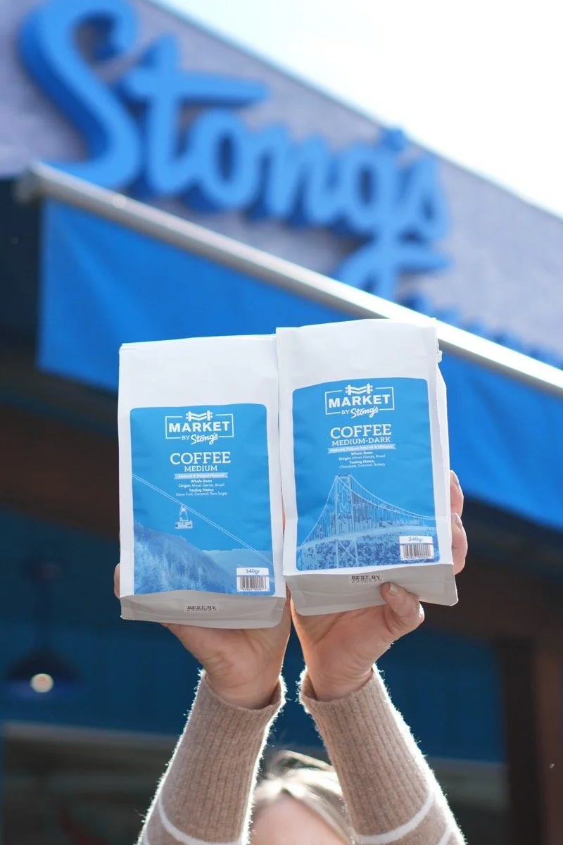 Hands holding two bags of medium dark coffee in front of a blue storefront with the word "Stong" visible in the background.