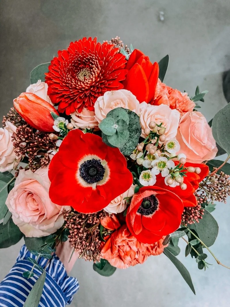 A colorful bouquet of flowers including red poppies, red gerbera daisies, and blush pink roses, with green foliage and white filler flowers.