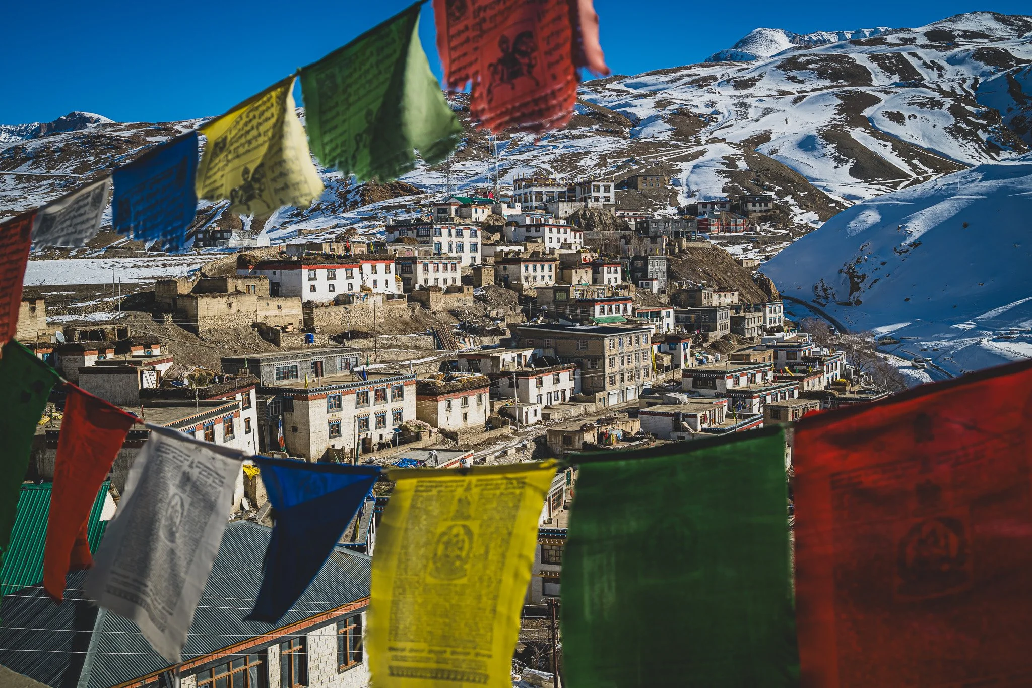 Bunte Gebetsfahnen vor Kibber mit verschneiten Bergen im Hintergrund.