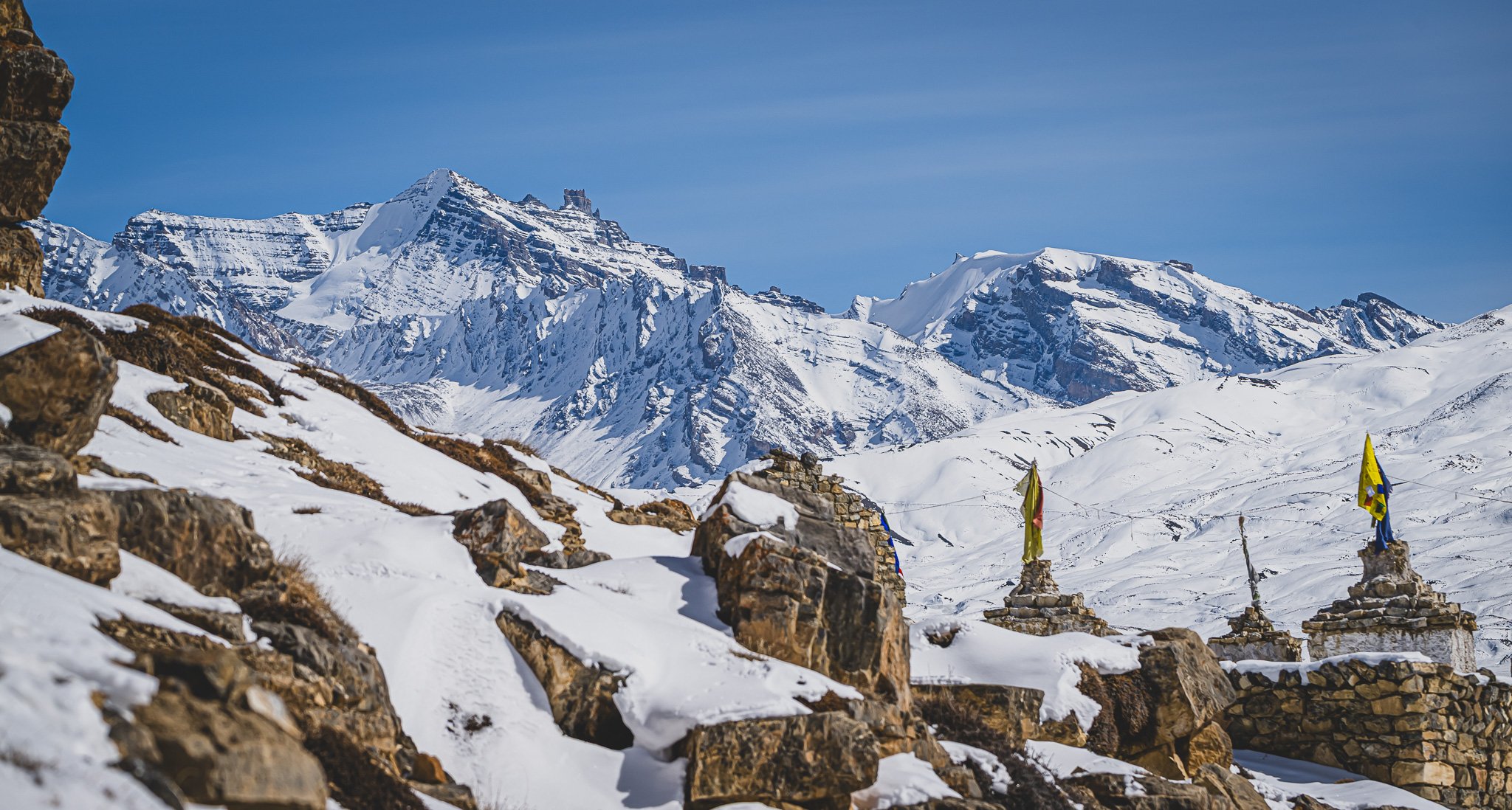 Schneebedeckte Berge und Steine, in der Mitte drei mit Flaggen bedeckte Steintürme in einer alpinen Landschaft.