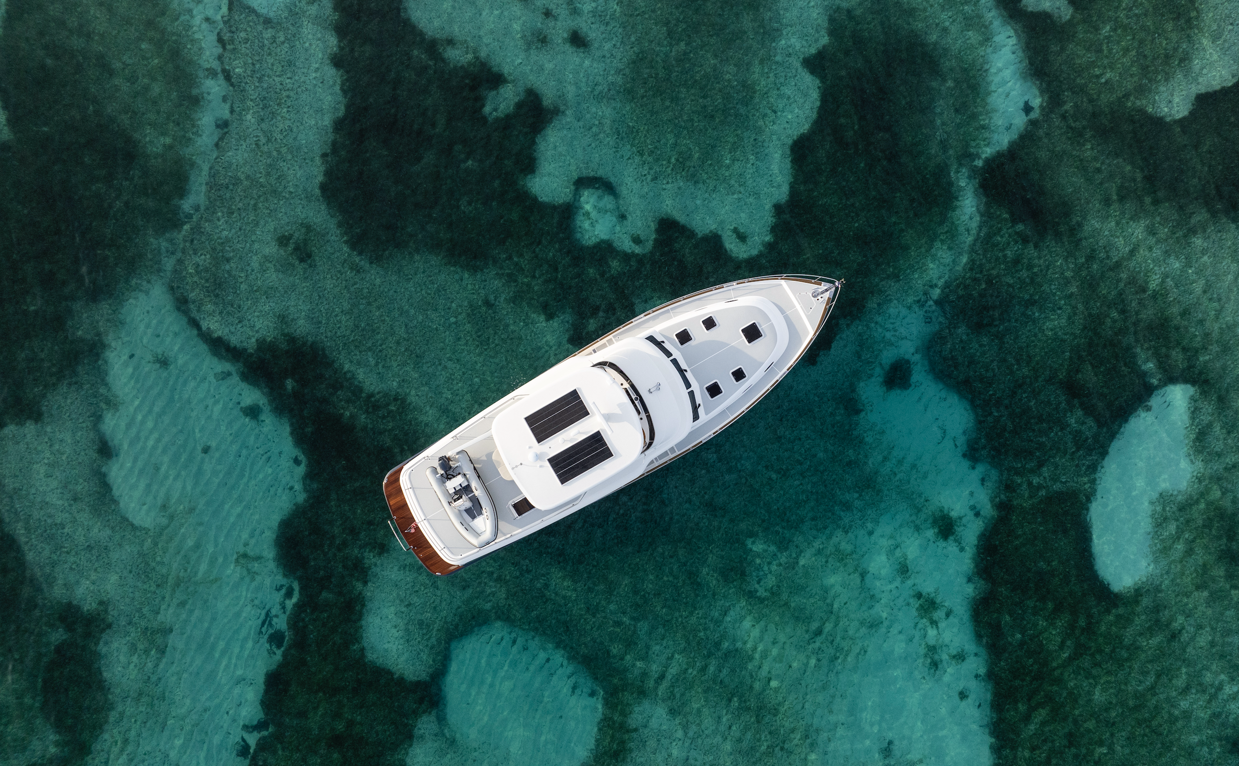 An aerial view of a white yacht sailing on clear turquoise water, with visible coral reefs underneath.
