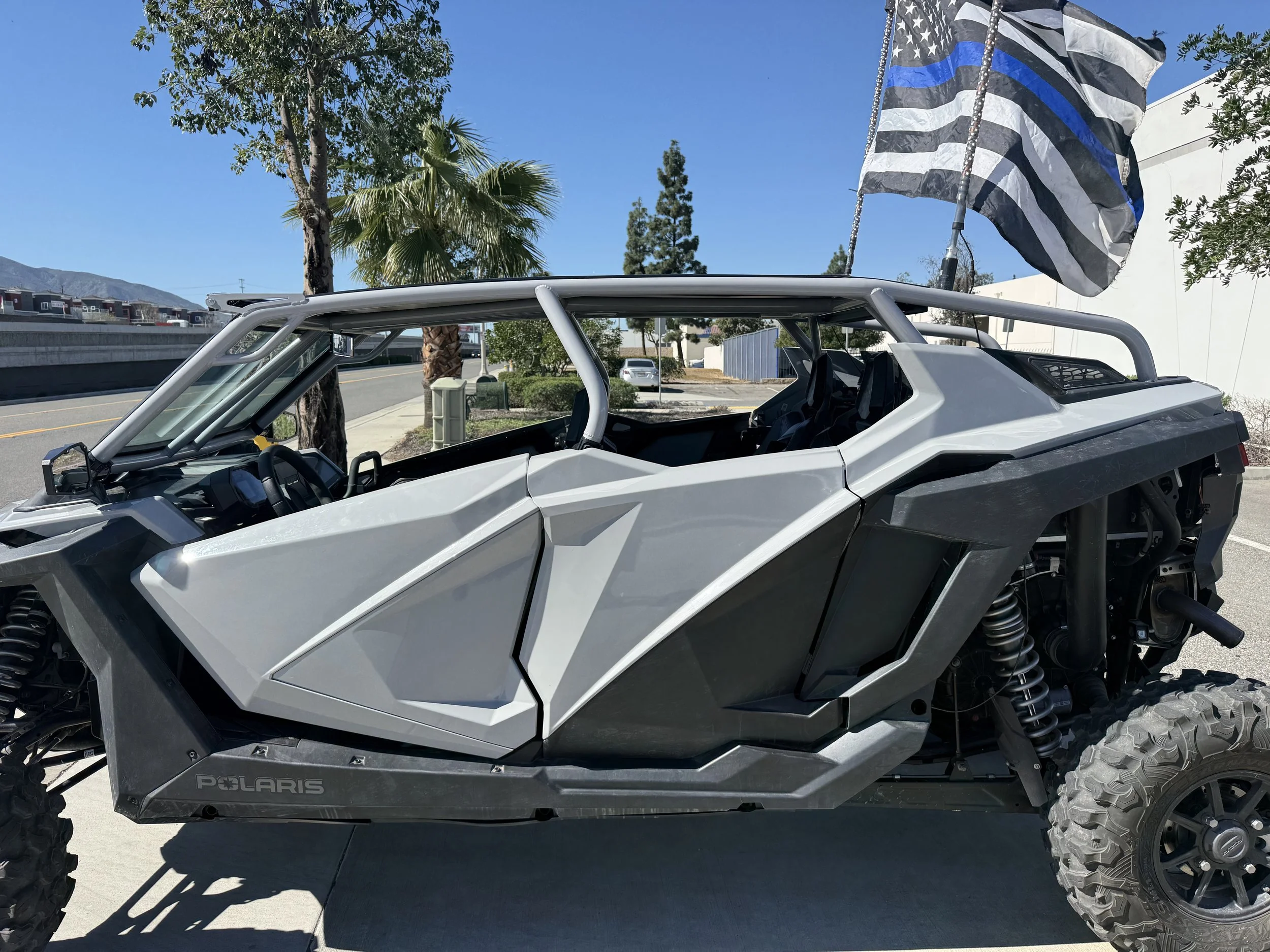 A white off-road Polaris vehicle with large tires, a black and white flag with a blue line, and a city street with trees and buildings in the background.