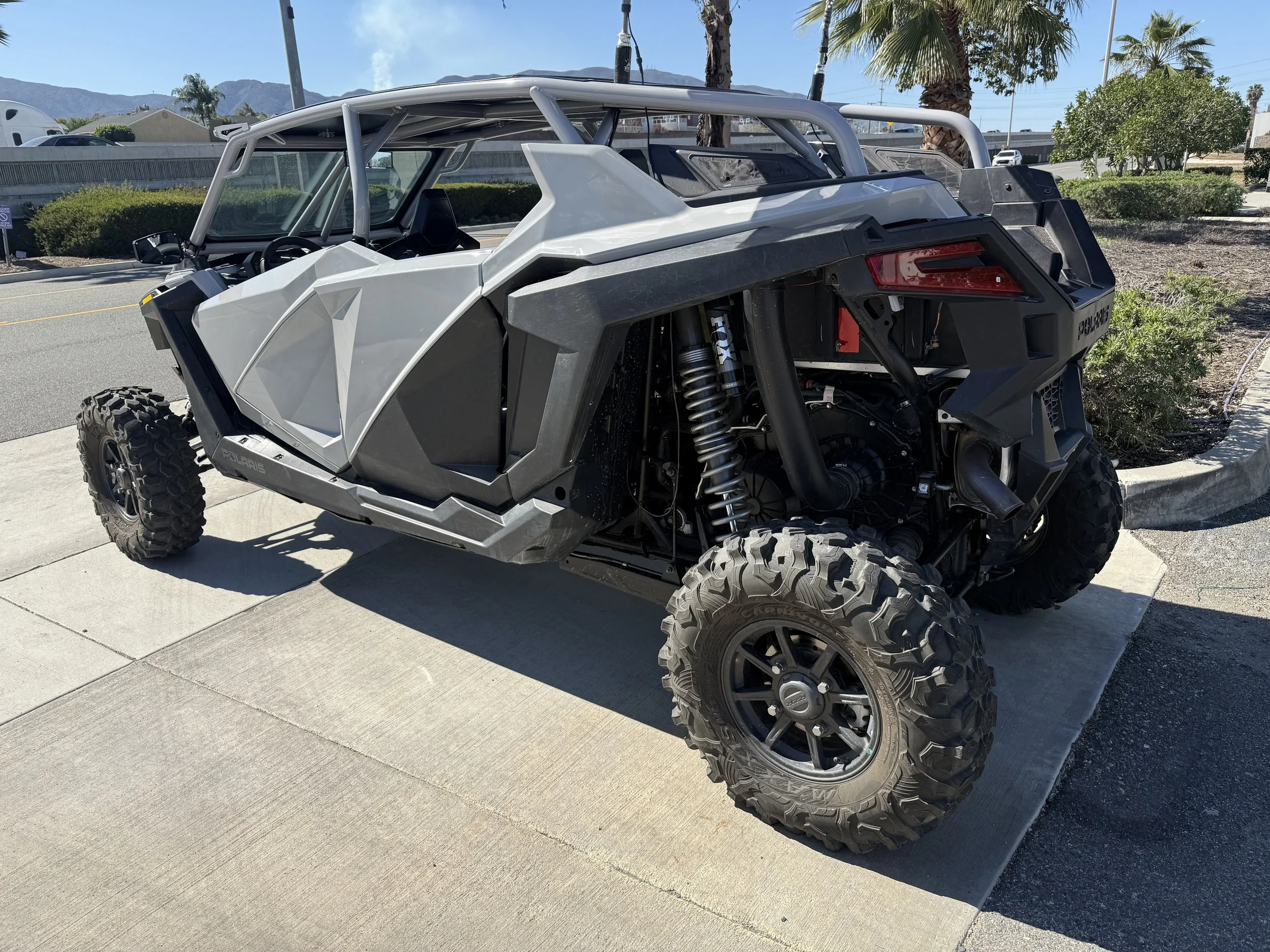 An off-road all-terrain vehicle (ATV) parked on a concrete sidewalk under clear sunny weather. The vehicle is black, gray, and white with large, rugged tires, exposed suspension, and a minimalist open design.