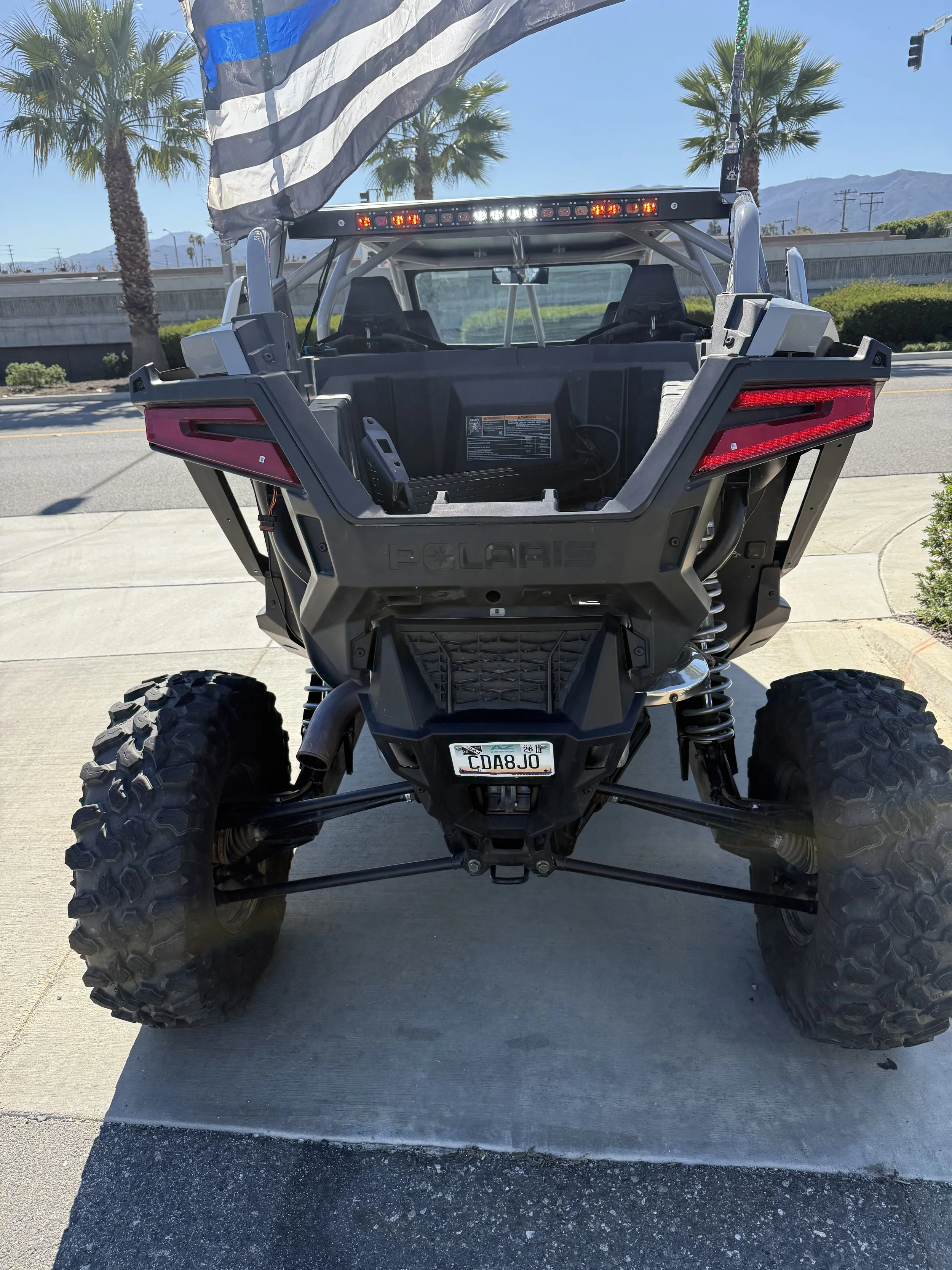 Front view of a black Polaris off-road vehicle with large tires, license plate 'CDA8JO', and an American flag attached, parked on a driveway with a clear blue sky, palm trees, and mountains in the background.