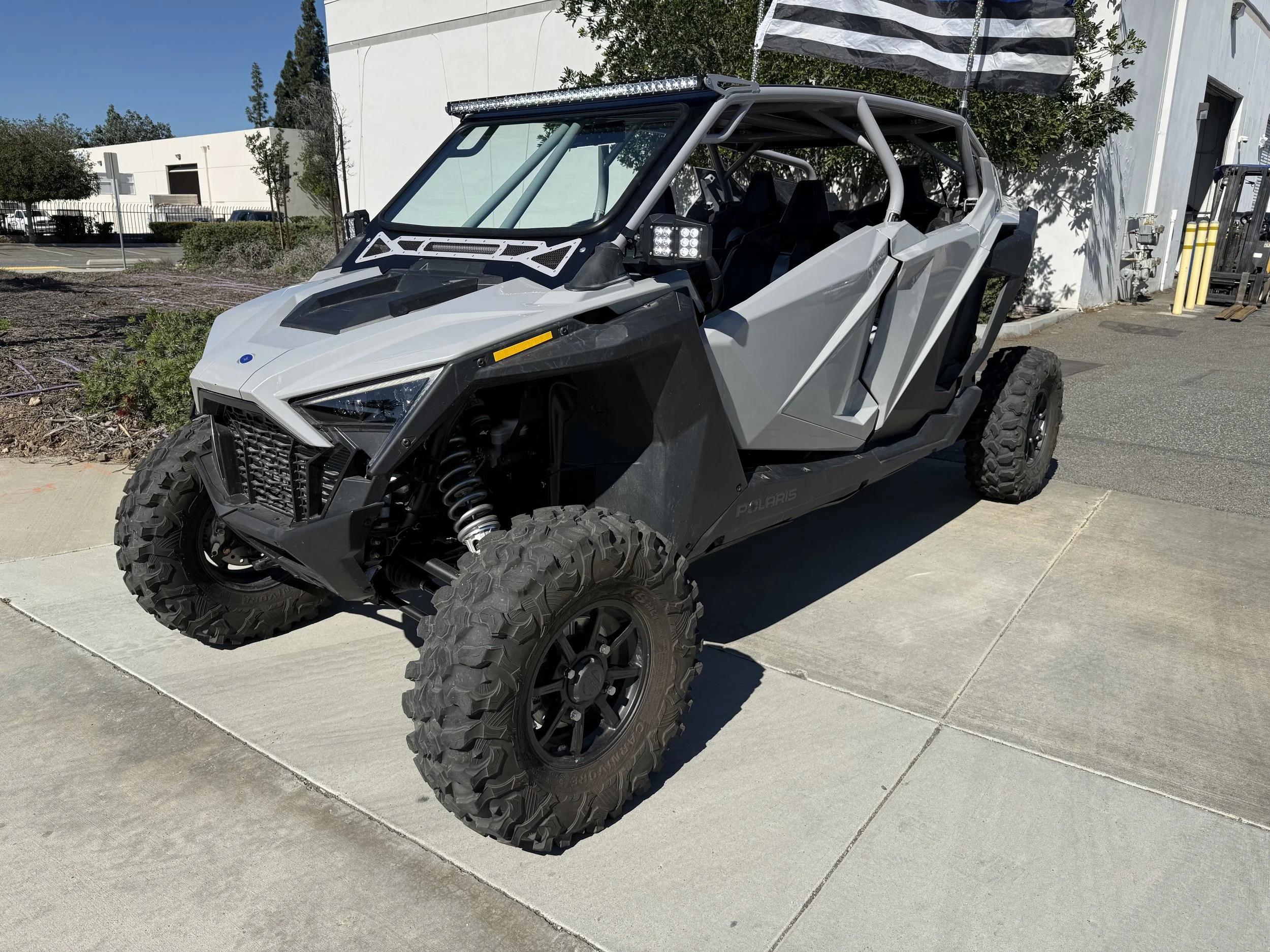Gray off-road utility vehicle with large tires, exposed suspension, and roll cage, parked on concrete pavement near white building and trees.