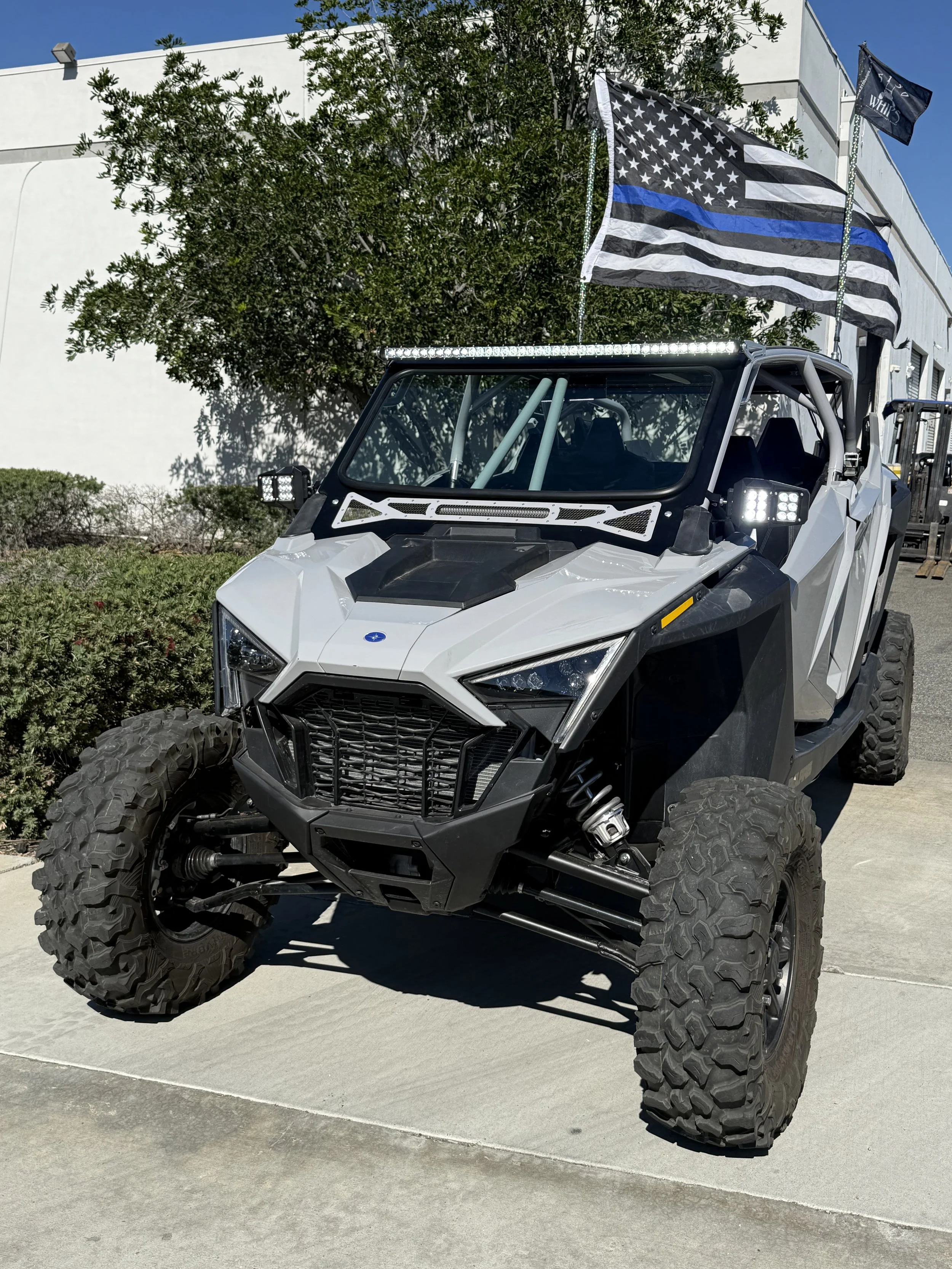 Off-road utility vehicle with a black and white color scheme, large rugged tires, and a flag with a black and white American flag with a blue stripe, parked outdoors on pavement with a building, bushes, and trees in the background.
