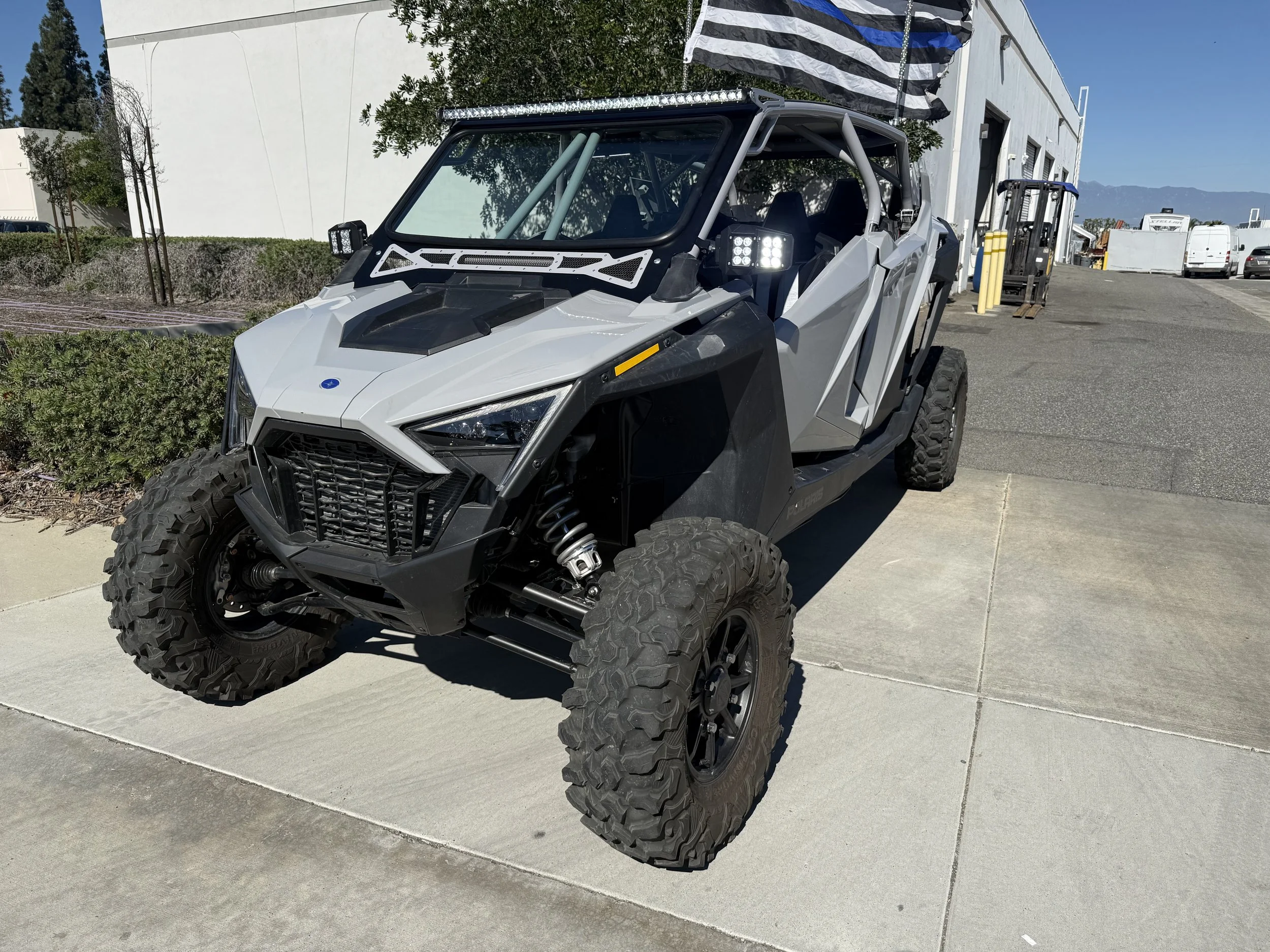 A futuristic off-road vehicle with large rugged tires, a gray and black angular body, and LED lights, parked outside a building with a palette of white, gray, and black colors, and a black and white flag flying overhead.