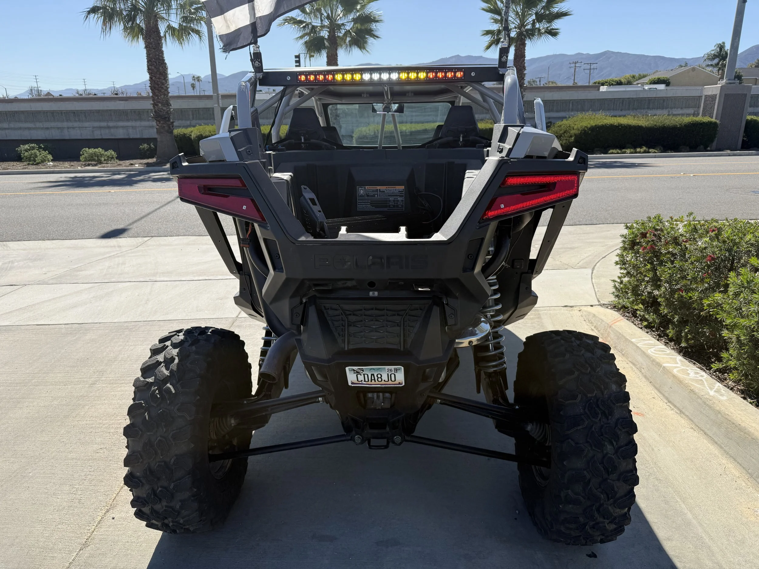 Rear view of a Polaris all-terrain vehicle (ATV) parked on a sidewalk with mountains, palm trees, and a crosswalk in the background.