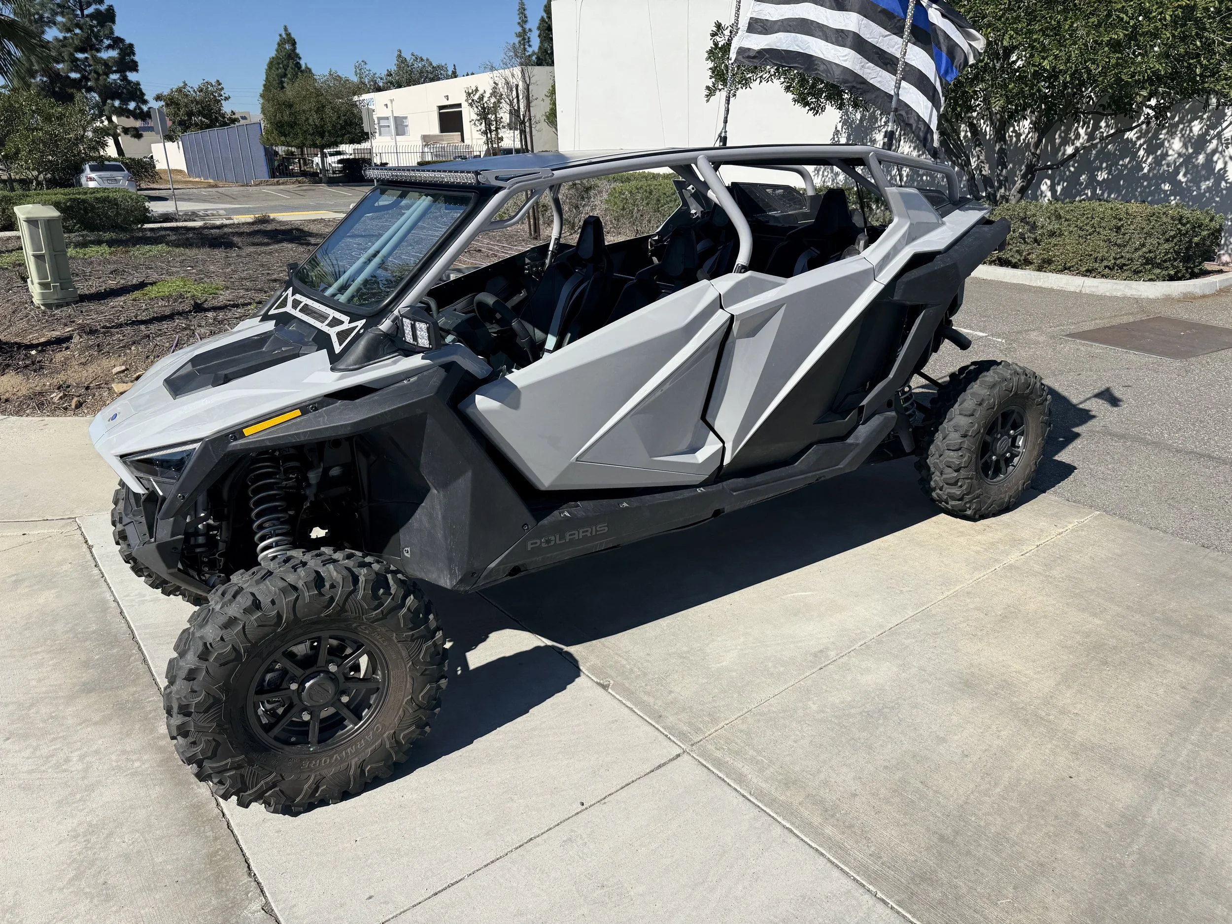 Gray Polaris off-road vehicle parked on a concrete sidewalk with a flag in the background and trees.