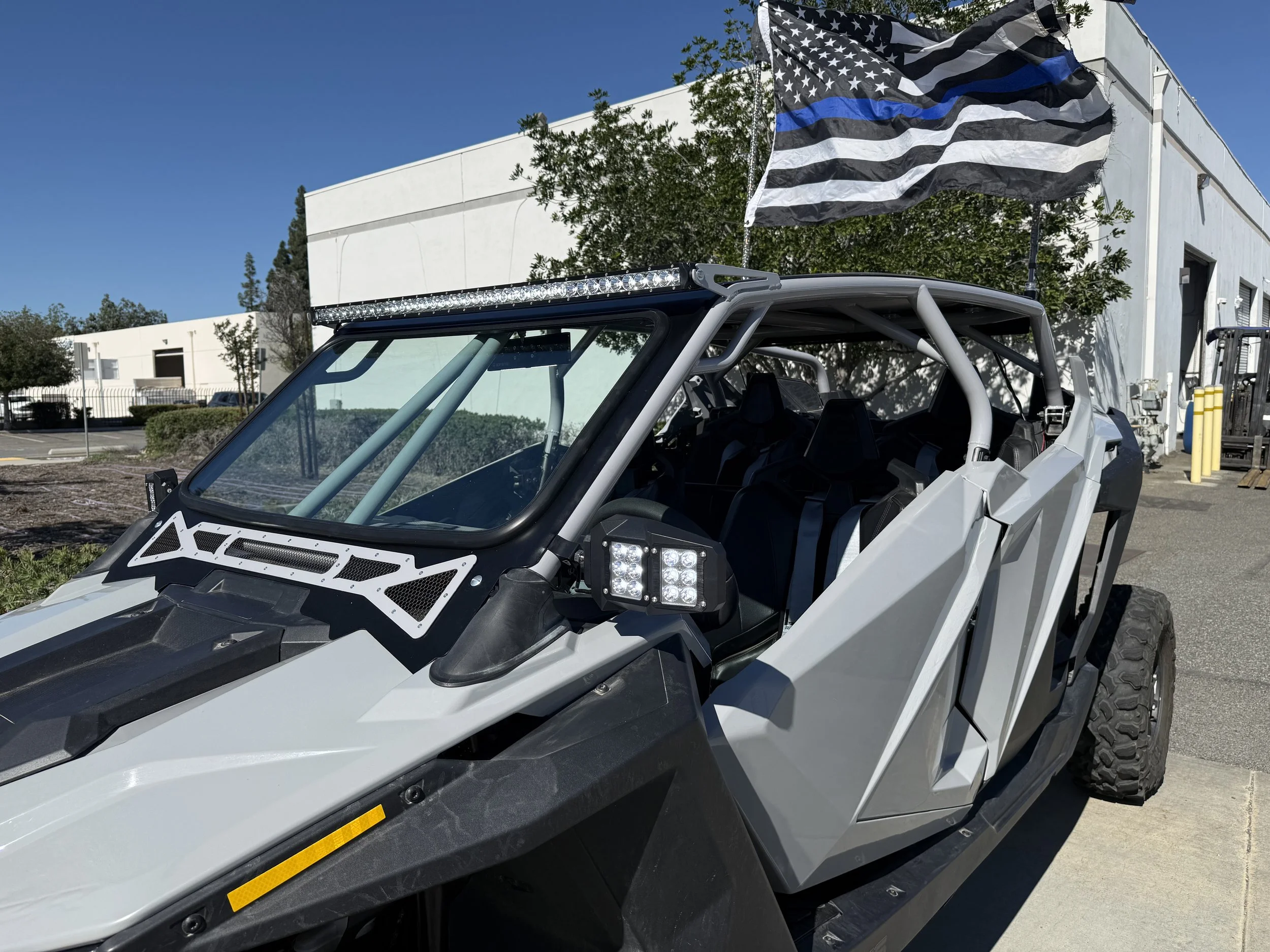 An off-road electric vehicle with an American flag with a black and blue stripe, parked outdoors next to a white building with trees and a blue sky in the background.