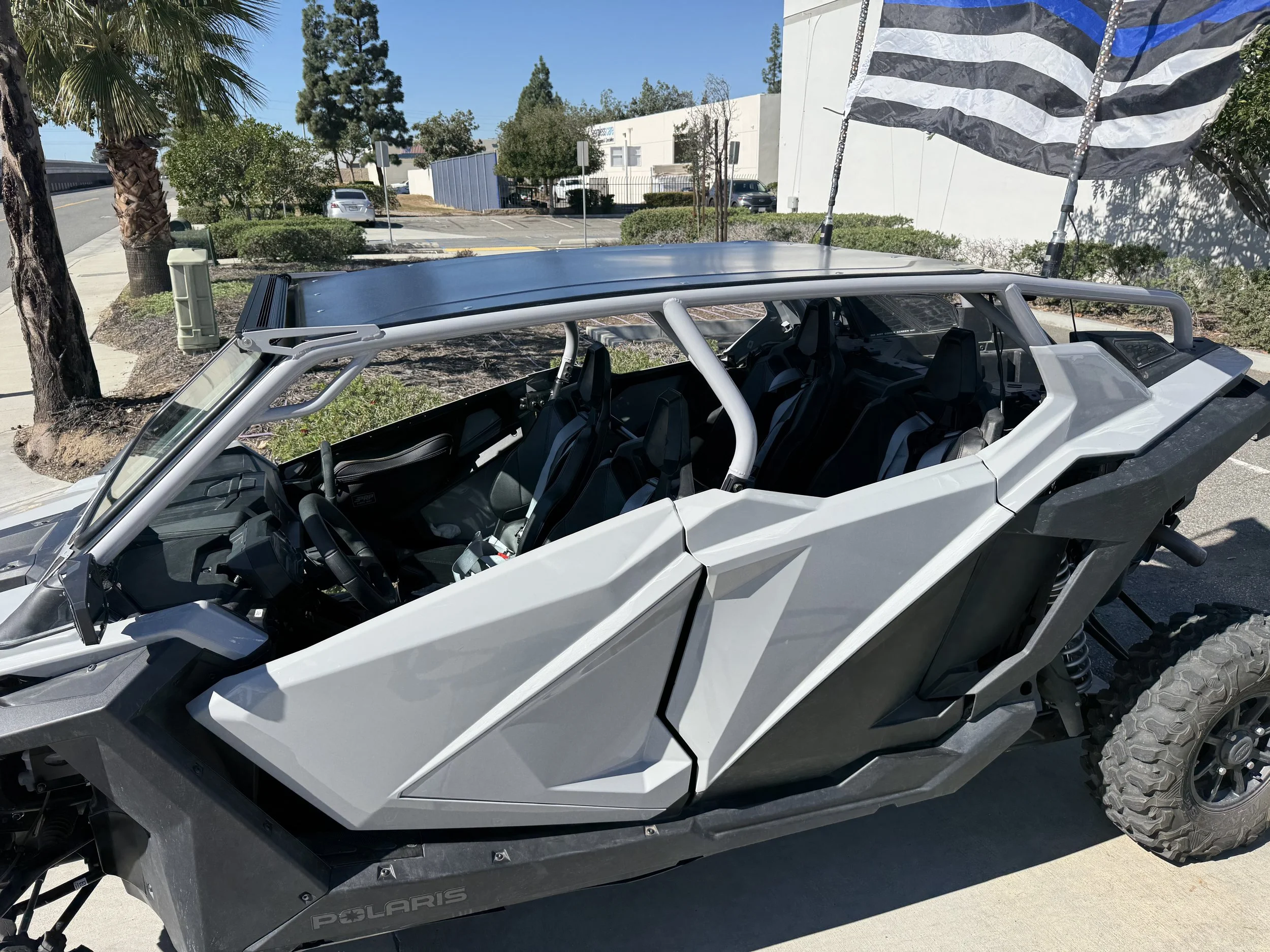 A gray Polaris off-road vehicle with six seats and an American flag mounted on the back, parked on a sidewalk with trees and buildings in the background.