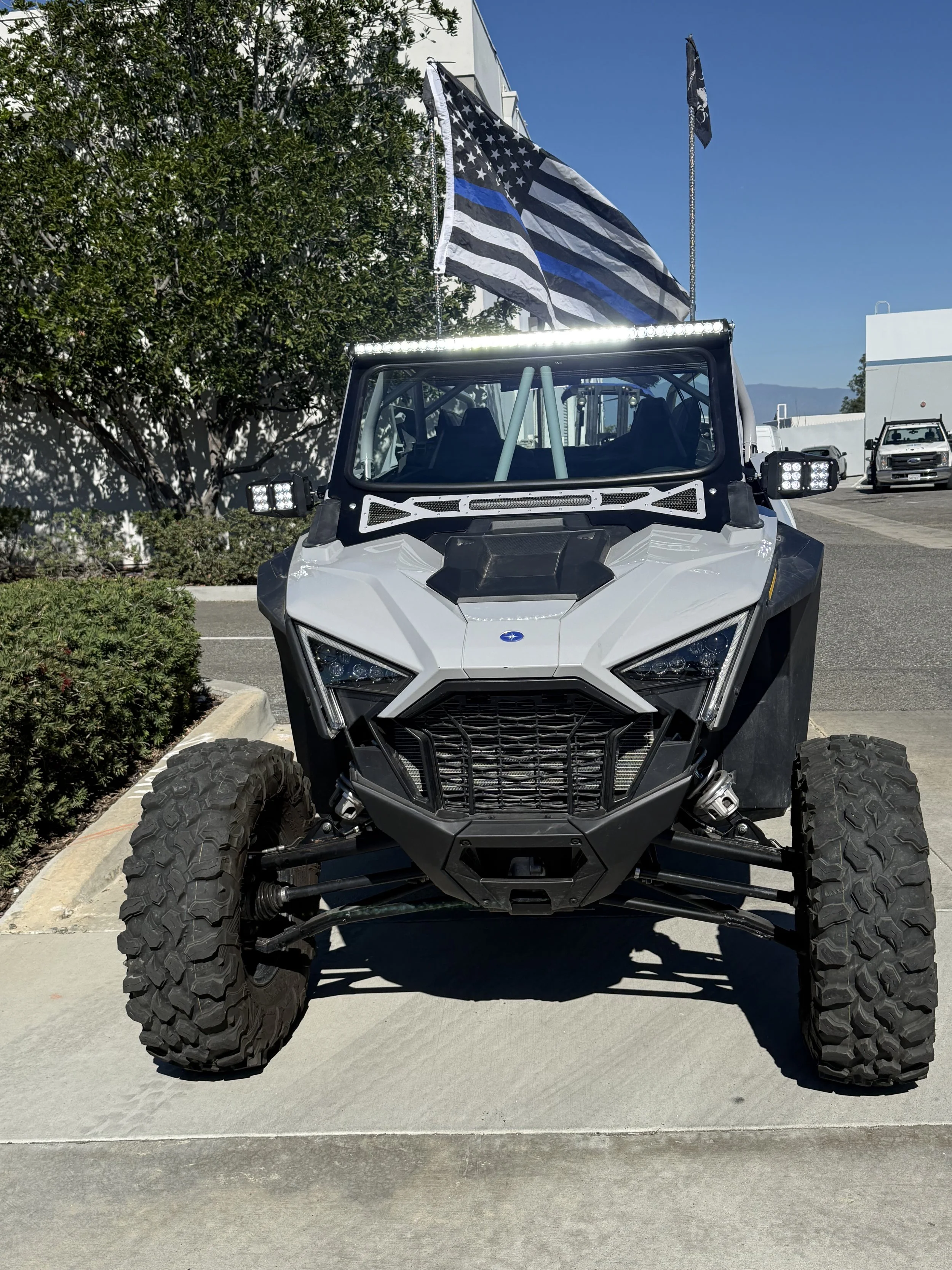 Off-road utility vehicle with two flags mounted on a pole, one American flag and a black and white flag, parked on a concrete sidewalk under a bright blue sky.