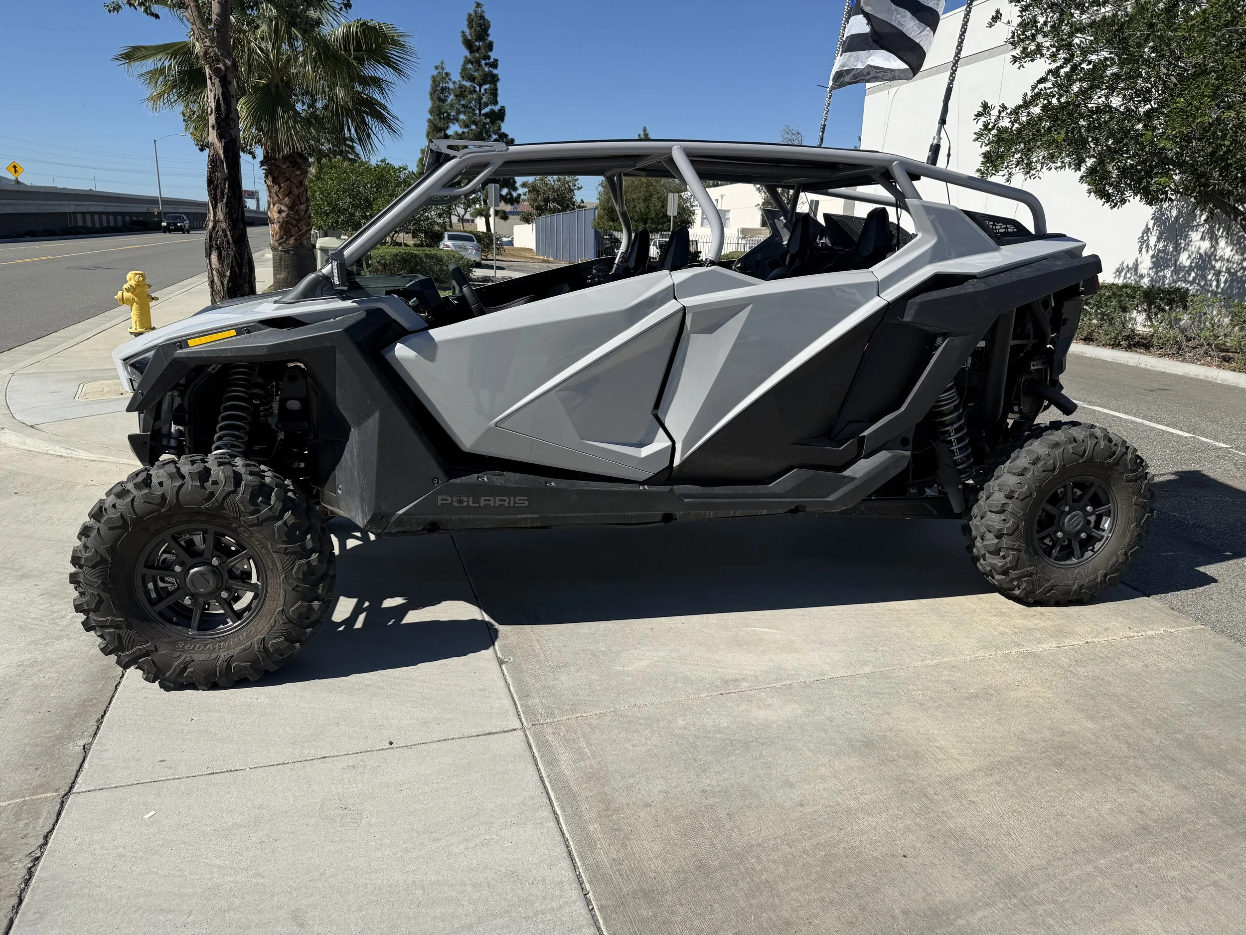 Grey off-road Polaris vehicle with large tires and open sides parked on the sidewalk on a sunny day.
