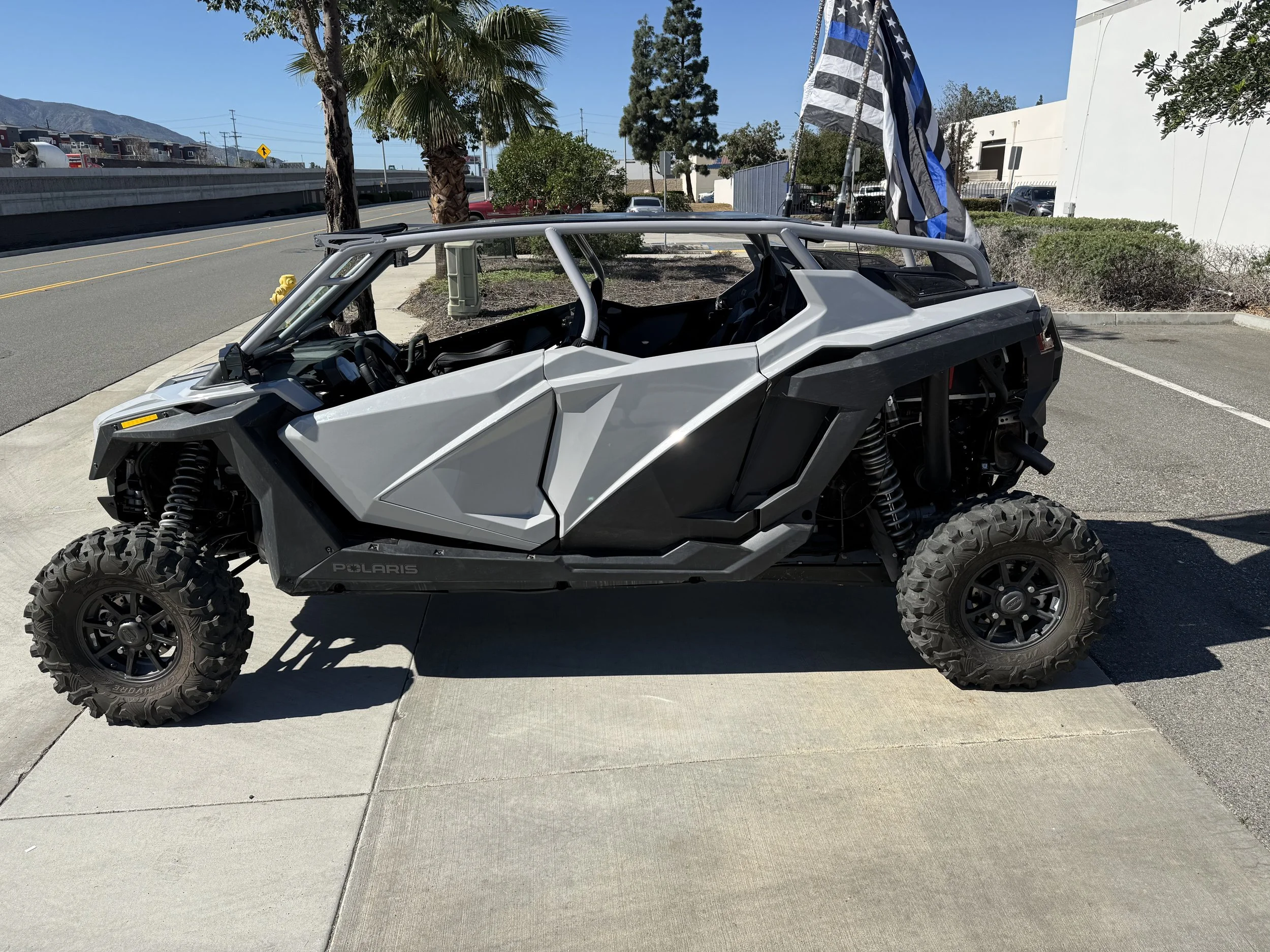 A Polaris off-road vehicle parked on a sidewalk with black and white striped blue flag attached to it, palm trees, a street, and buildings in the background.