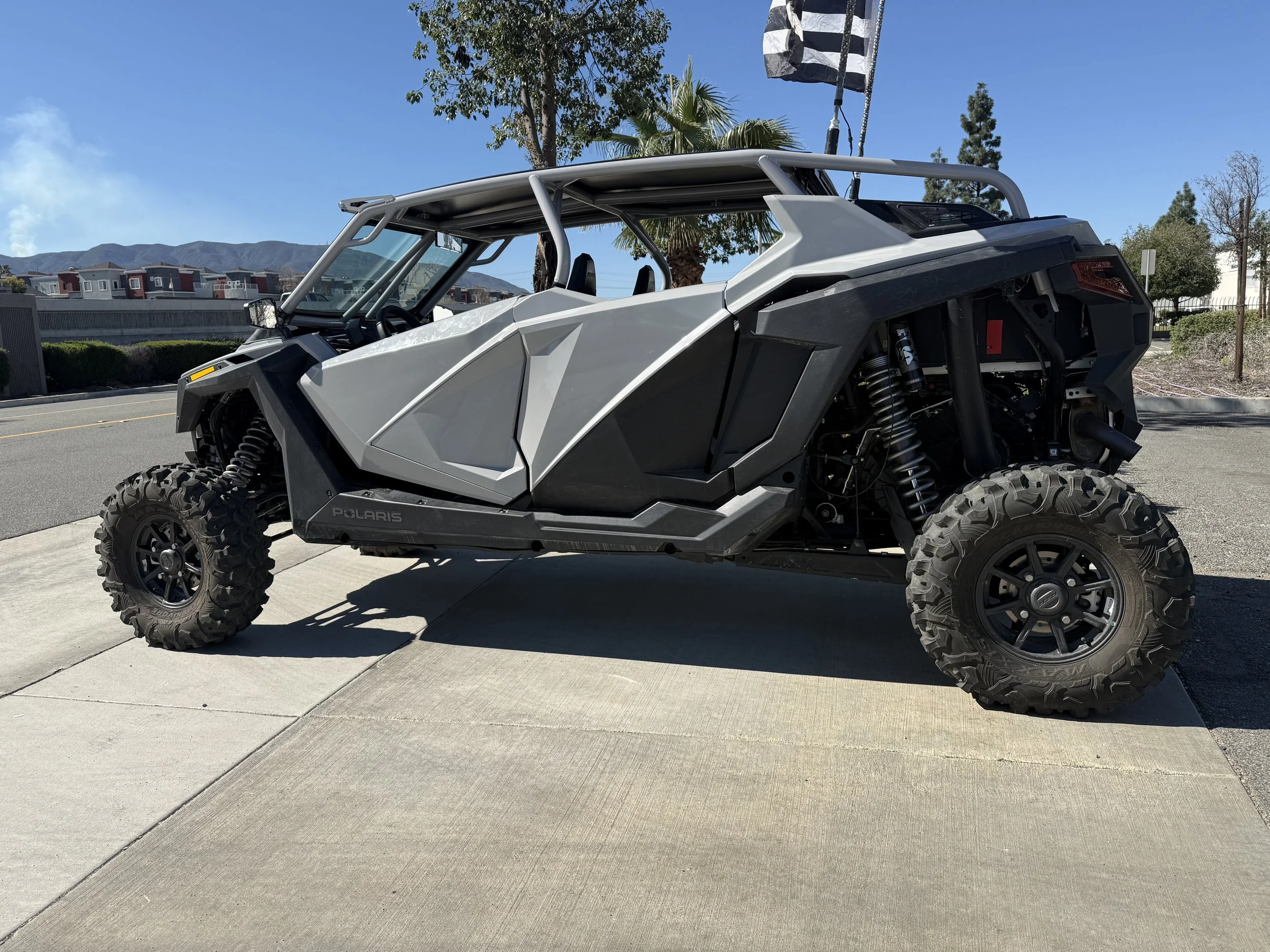 An off-road Polaris vehicle with rugged tires parked on a sidewalk under a clear sky, with trees and buildings in the background.