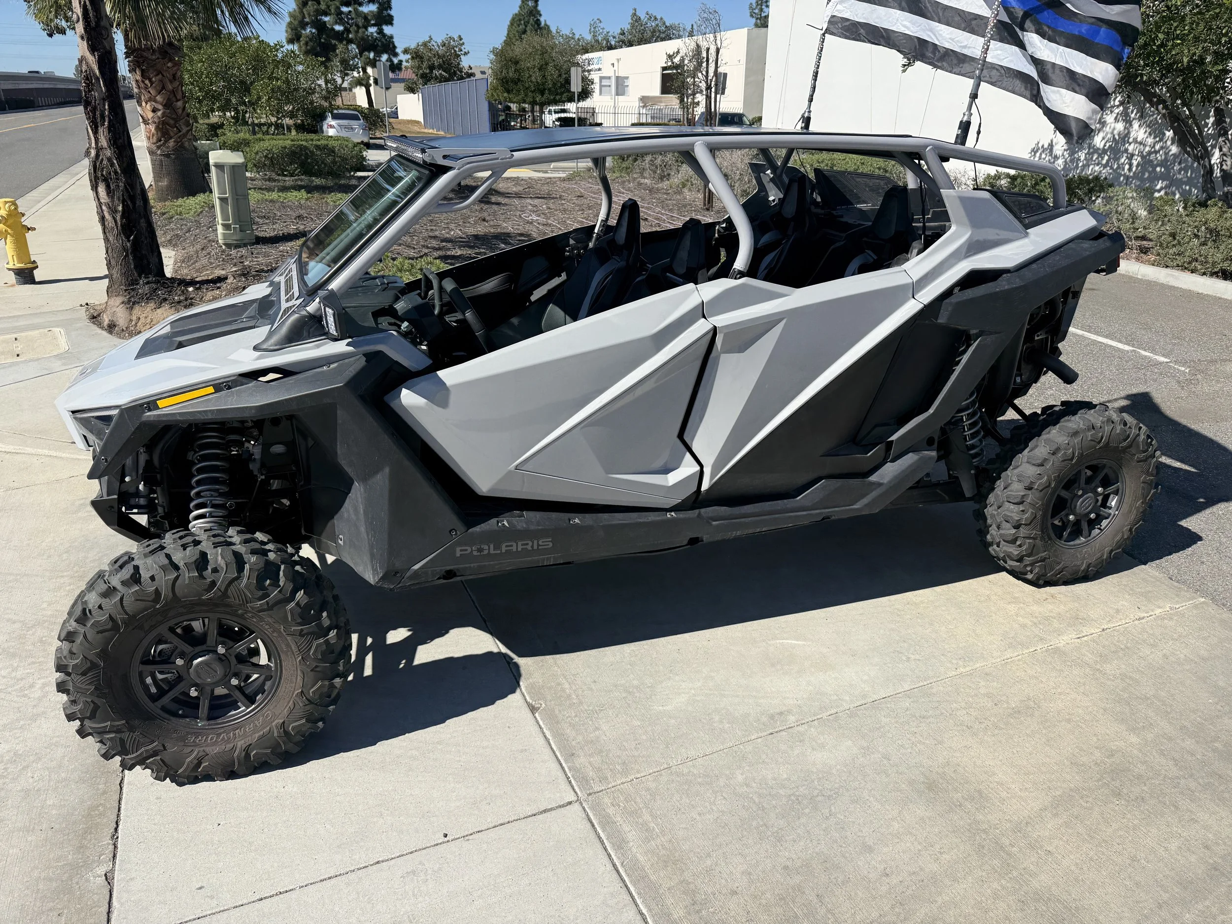 Gray Polaris off-road utility vehicle parked on concrete sidewalk near a street, with trees, bushes, and buildings in the background.