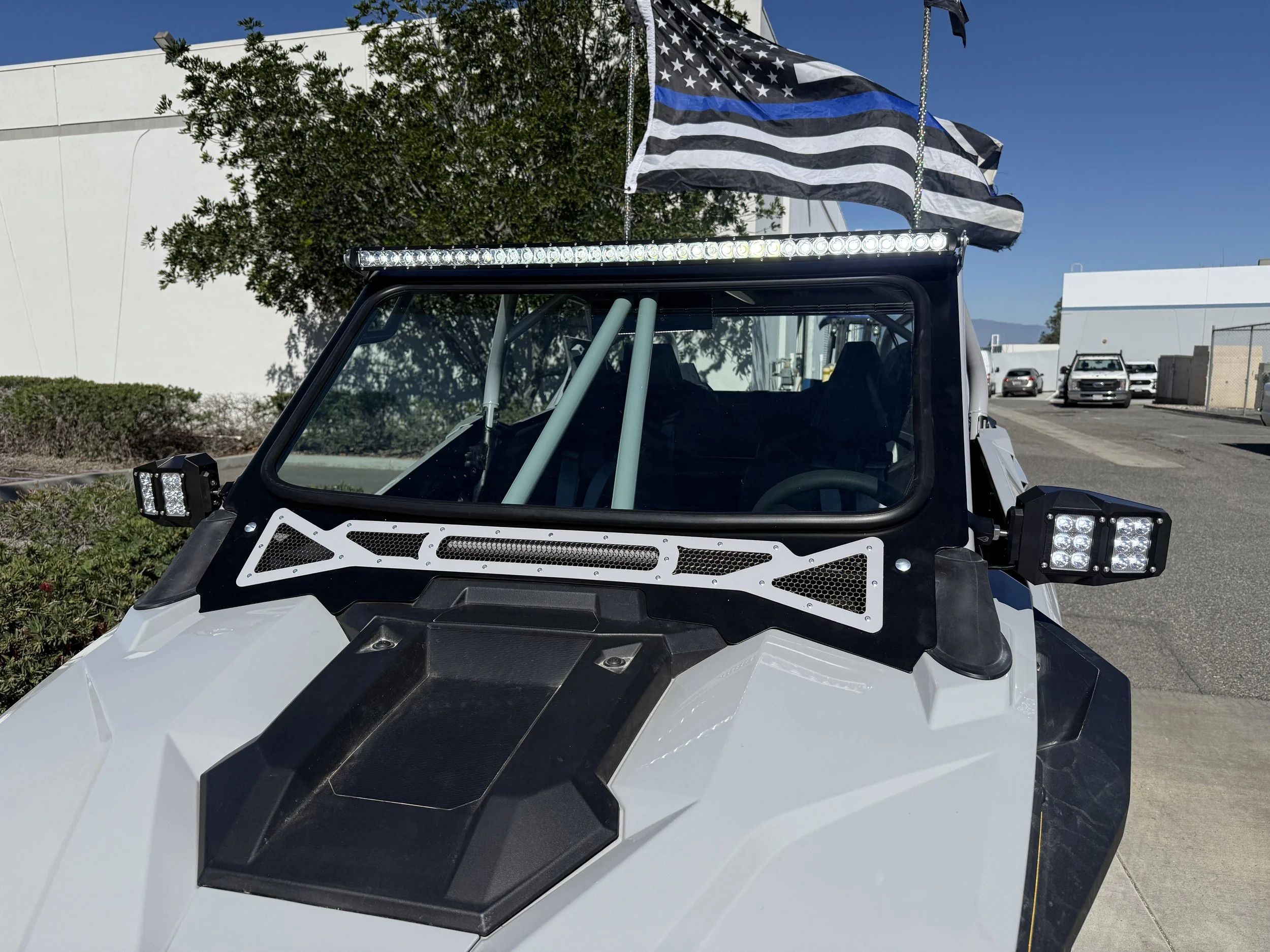 A white off-road vehicle with black accents parked outside, featuring a black and blue striped American flag flag on the roof, LED lights on the sides and top, and a roll cage inside.