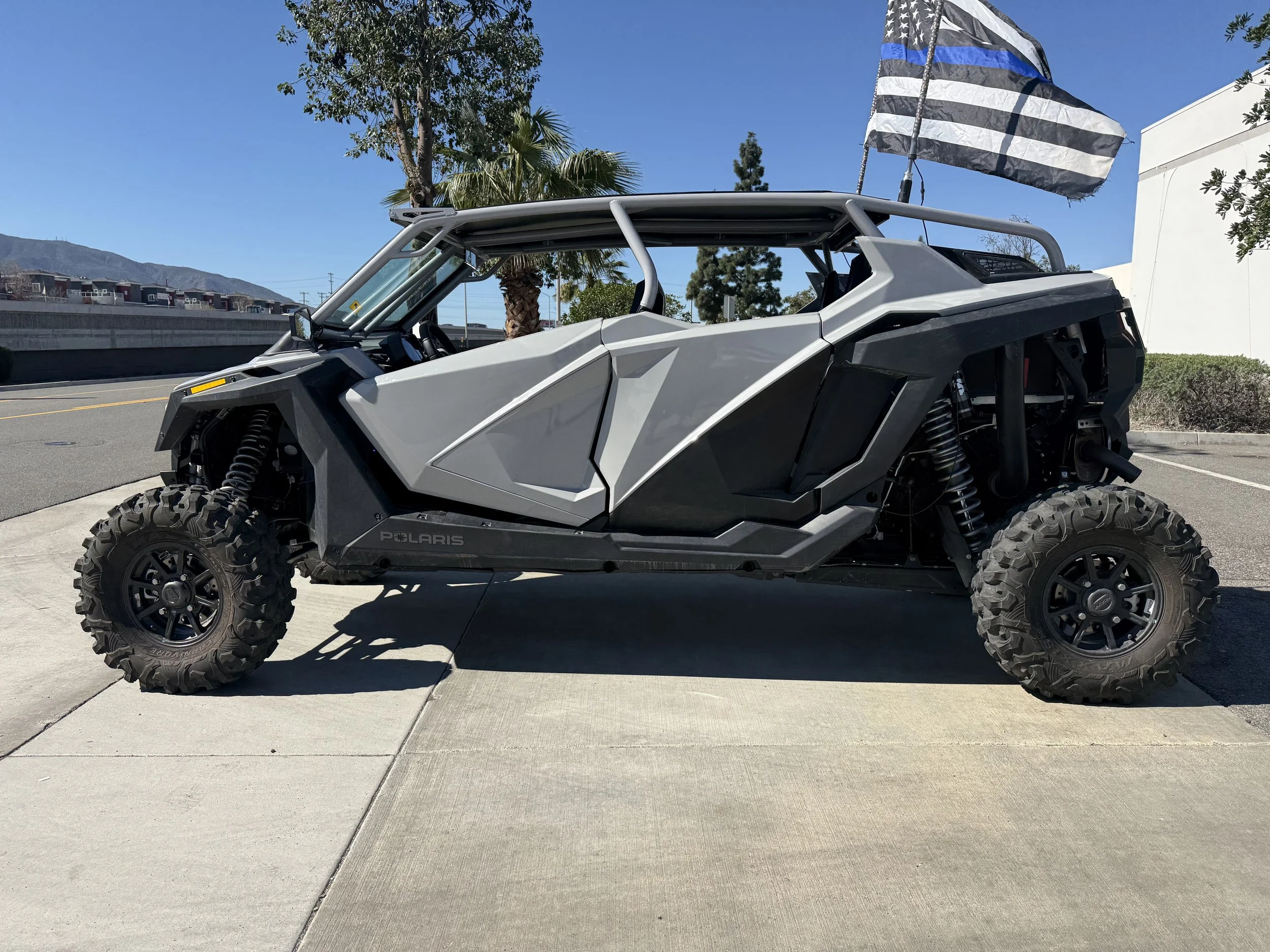 A gray off-road utility vehicle with large knobby tires and a blue and white striped flag attached, parked on a sidewalk in a sunny area with trees and a partially white building in the background.
