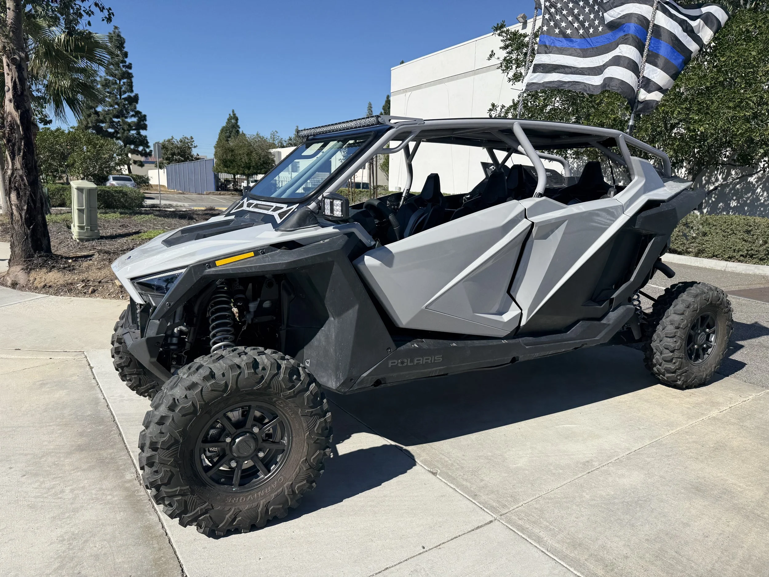 Gray Polaris off-road vehicle with large tires, parked on concrete sidewalk, with American flag in background.