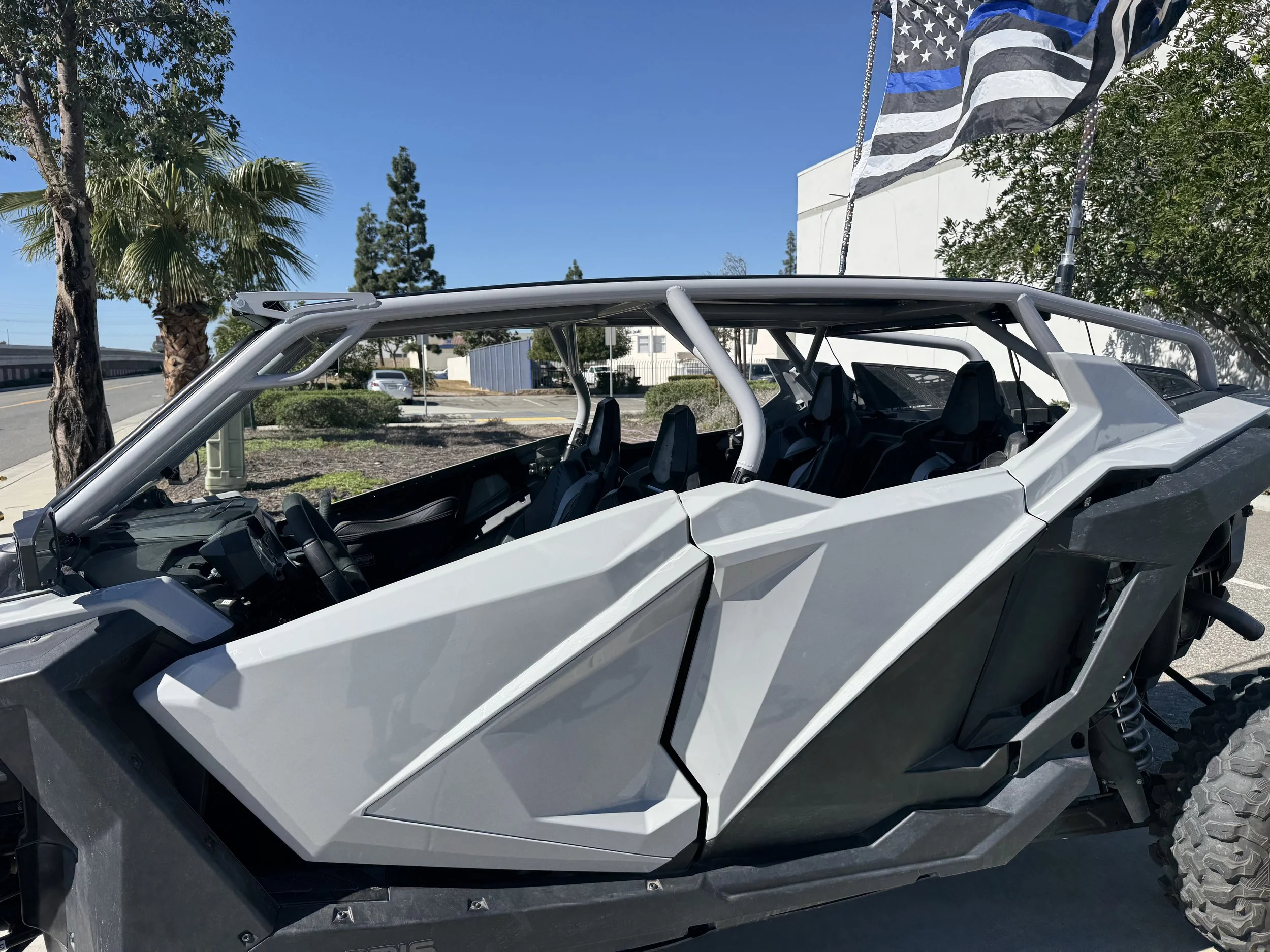 Off-road utility vehicle with a roll cage, black interior, and large rugged tires, parked outdoors near trees and a building under a clear blue sky, with a black and white American flag in the background.