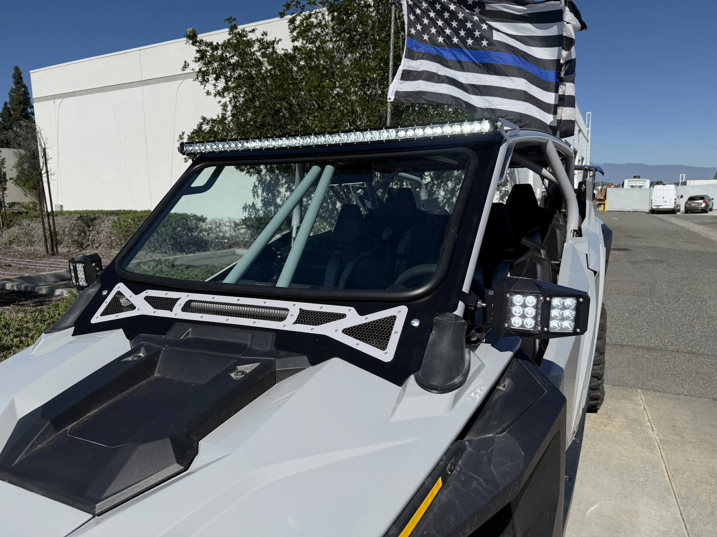 Close-up of a white off-road vehicle with a black roof, LED light bar, LED headlights, and a black and white checkered flag in the background, parked on a street with trees and buildings.