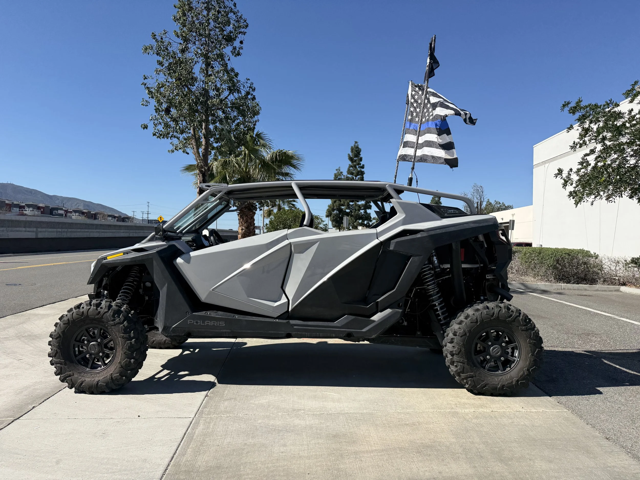 A gray Polaris off-road vehicle with large tires parked on a sidewalk, with blue sky, trees, and flags, including a black and white American flag, waving in the background.