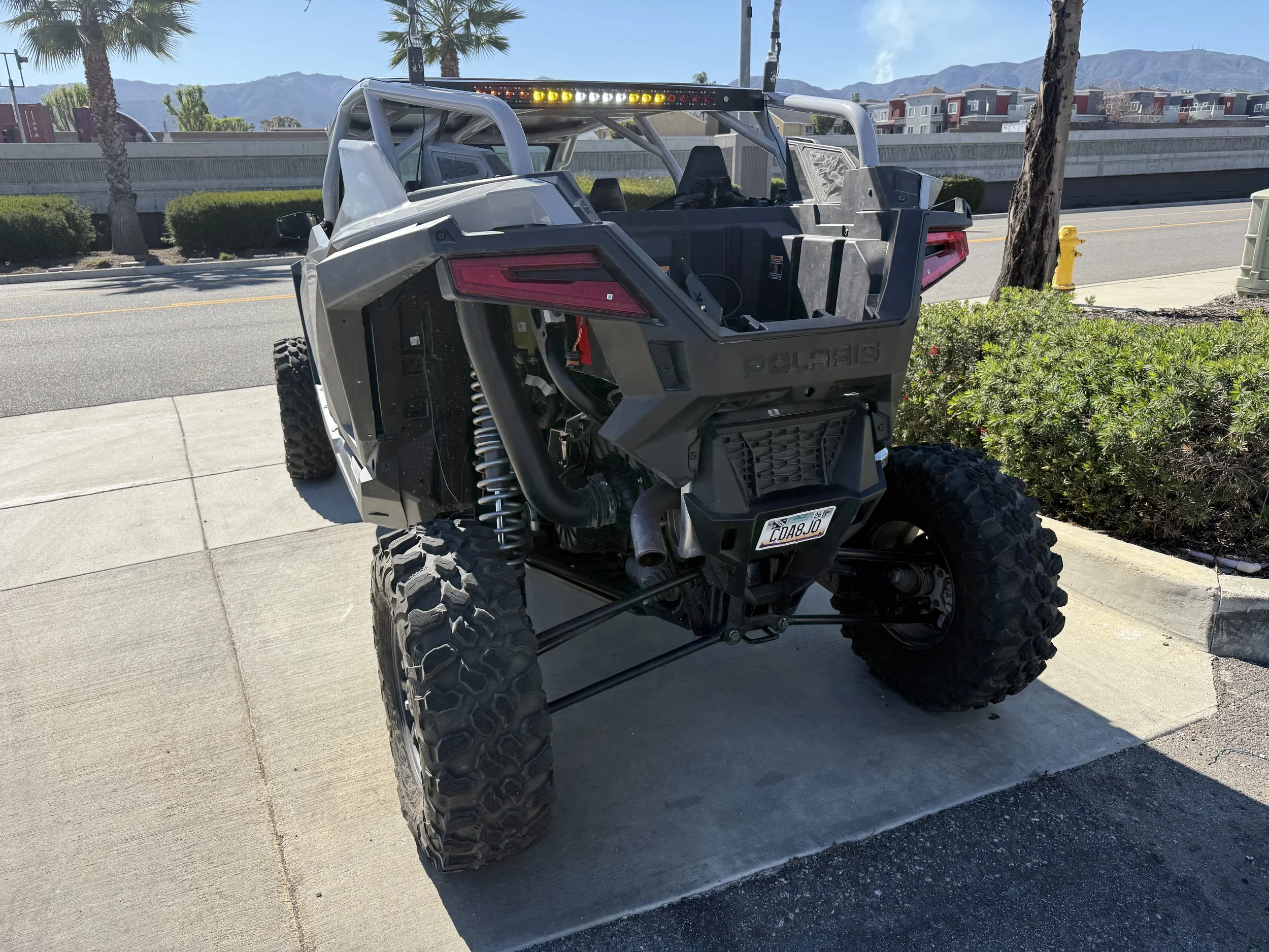 A black and gray Polaris off-road vehicle with large rugged tires, parked on a sidewalk near some bushes.