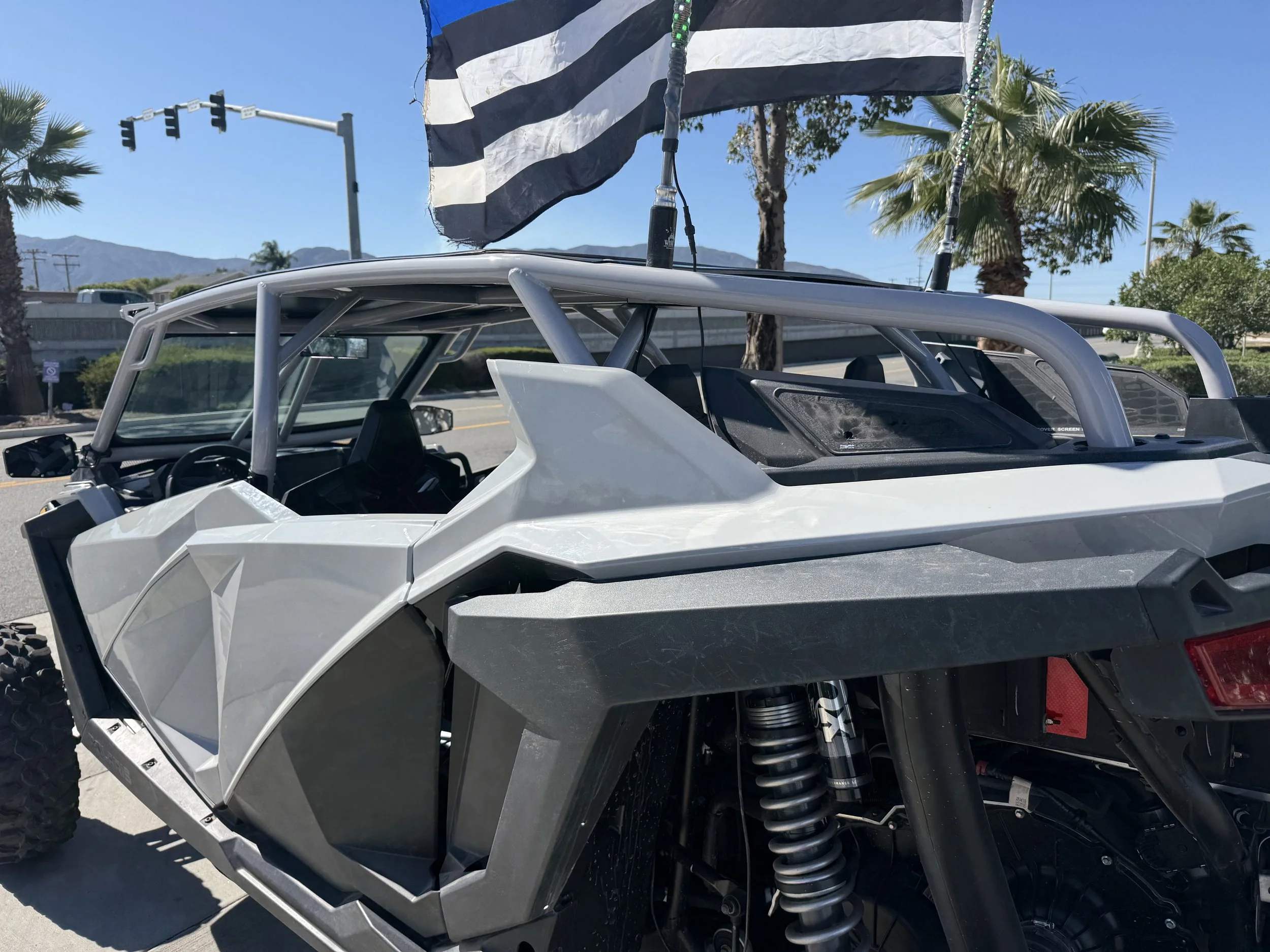 A Polaris off-road vehicle with a black and white striped flag flying above it, parked on a sunny street with palm trees, traffic lights, and mountains in the background.