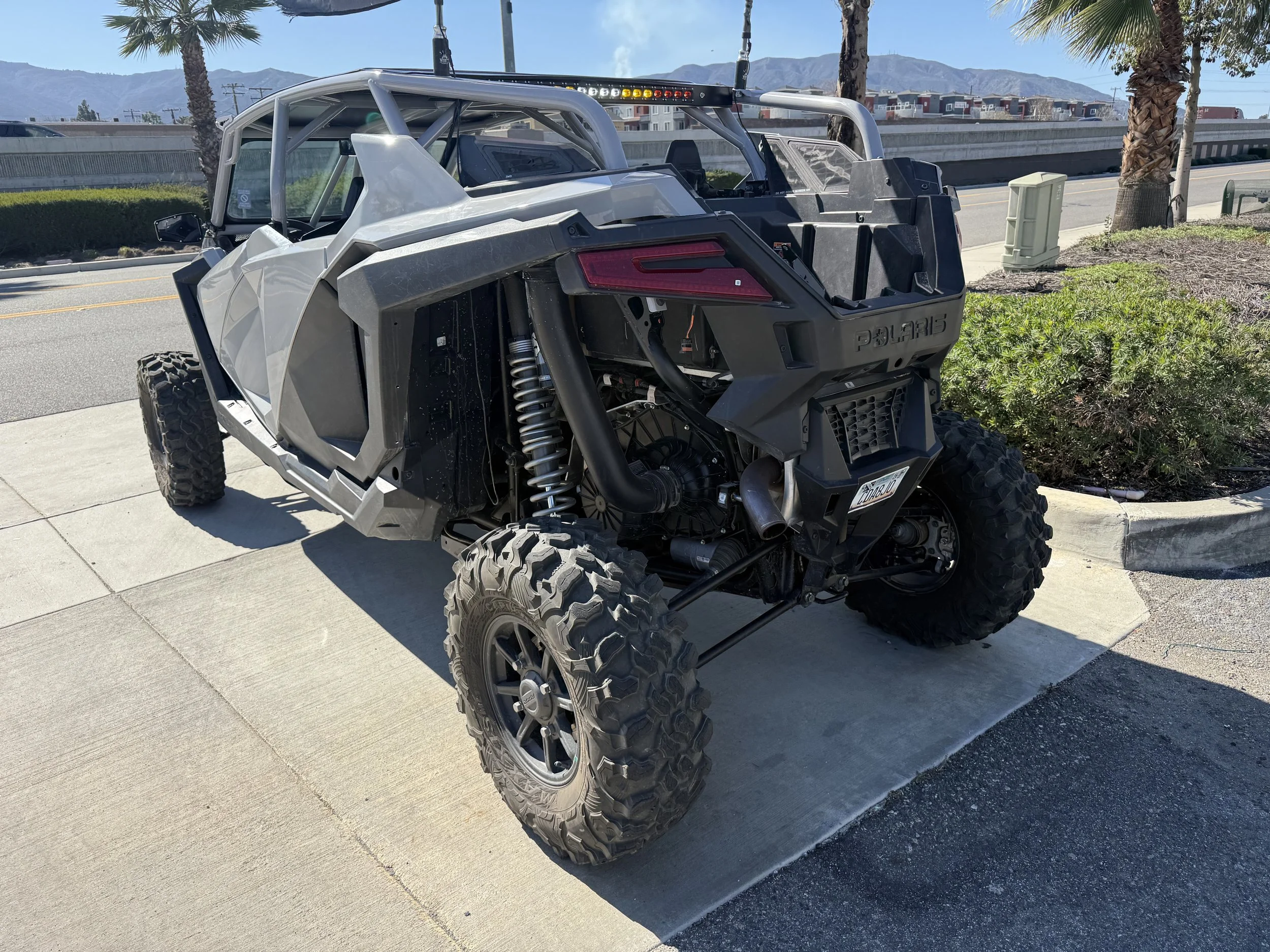 A black and gray off-road Polaris vehicle parked on a sidewalk with mountains and a highway in the background.