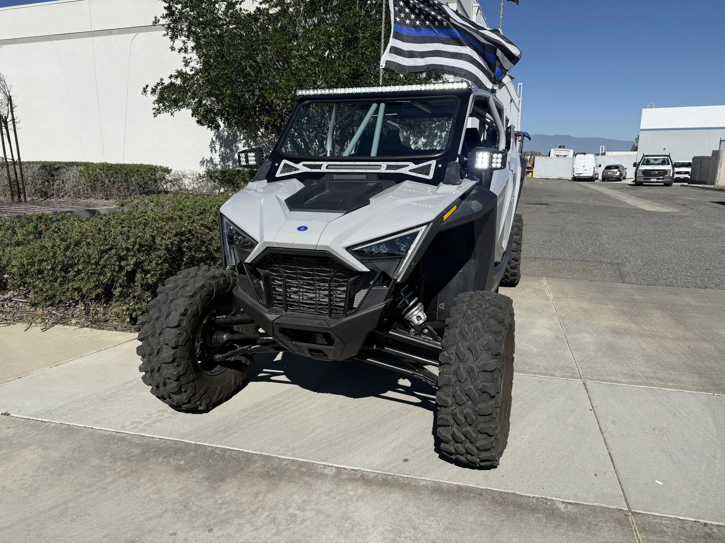 A white and black off-road utility vehicle with large tires, a roll cage, and lights, parked on a concrete sidewalk with an American flag attached on top.
