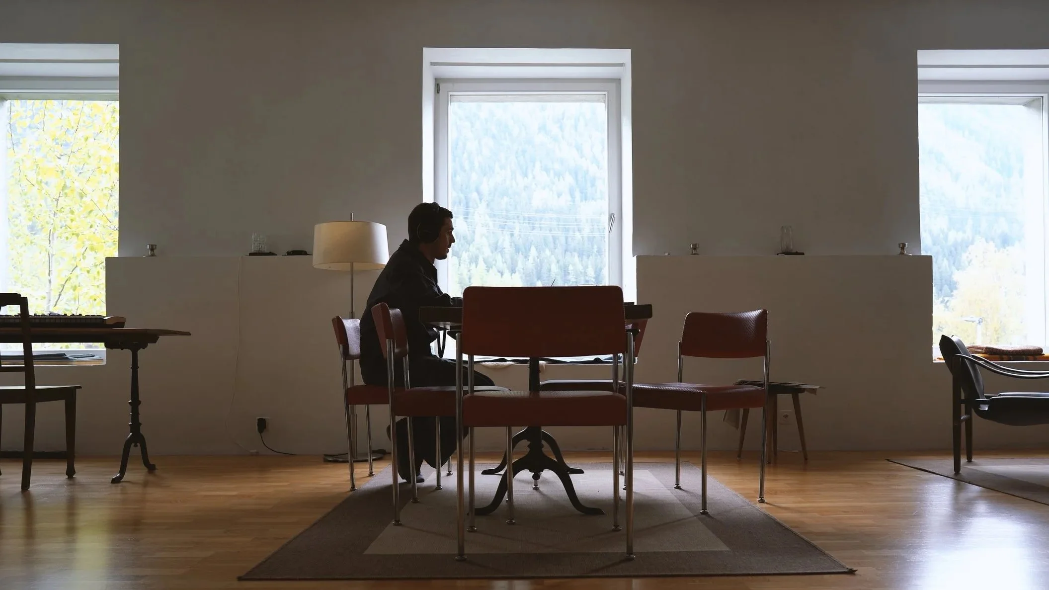 A person sitting at a table using a laptop in a room with large windows showing a view of trees and mountains outside.