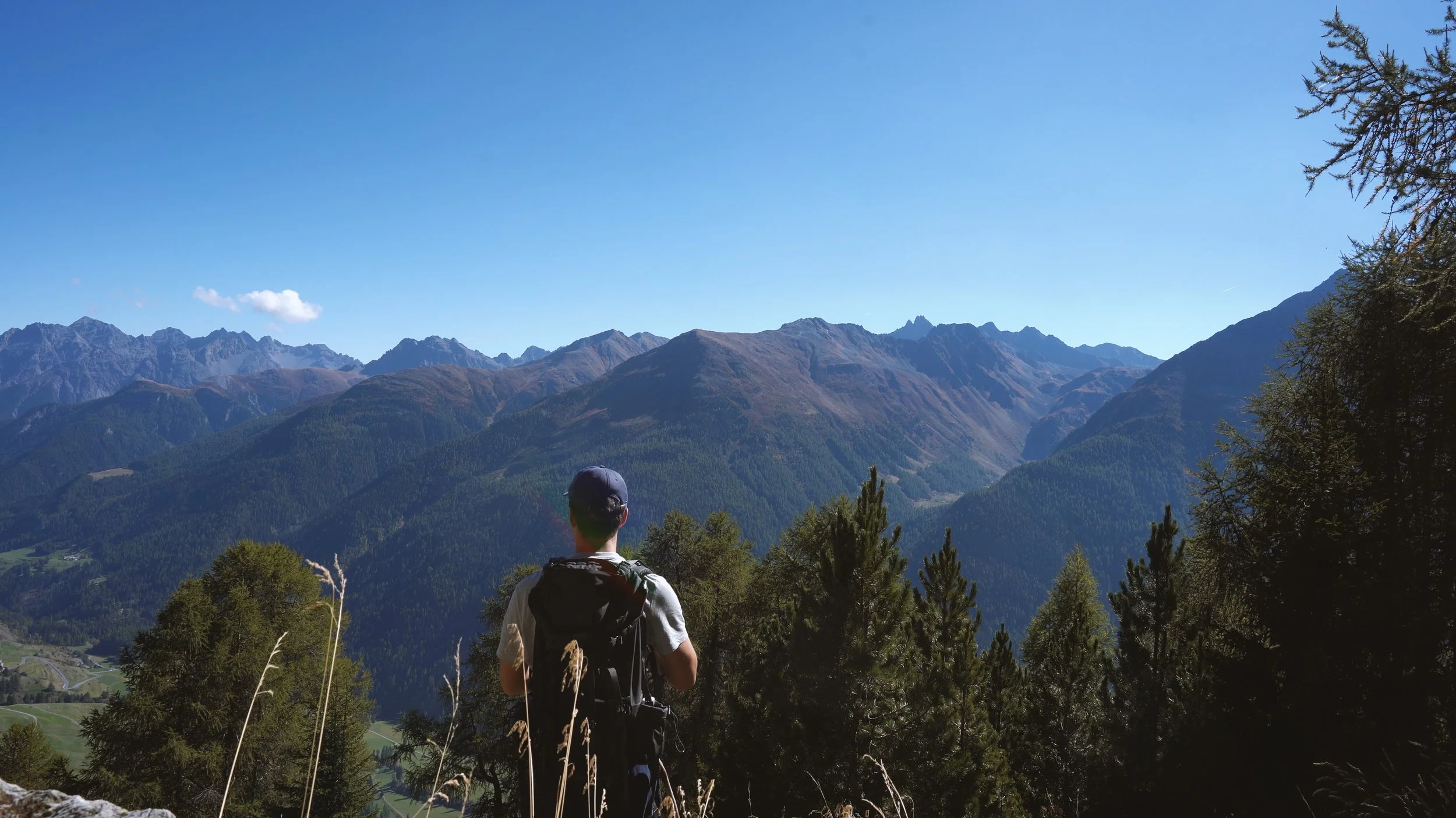 A person with a backpack and cap hiking in a mountainous area with a clear blue sky, green forests, and rugged mountain peaks in the background.