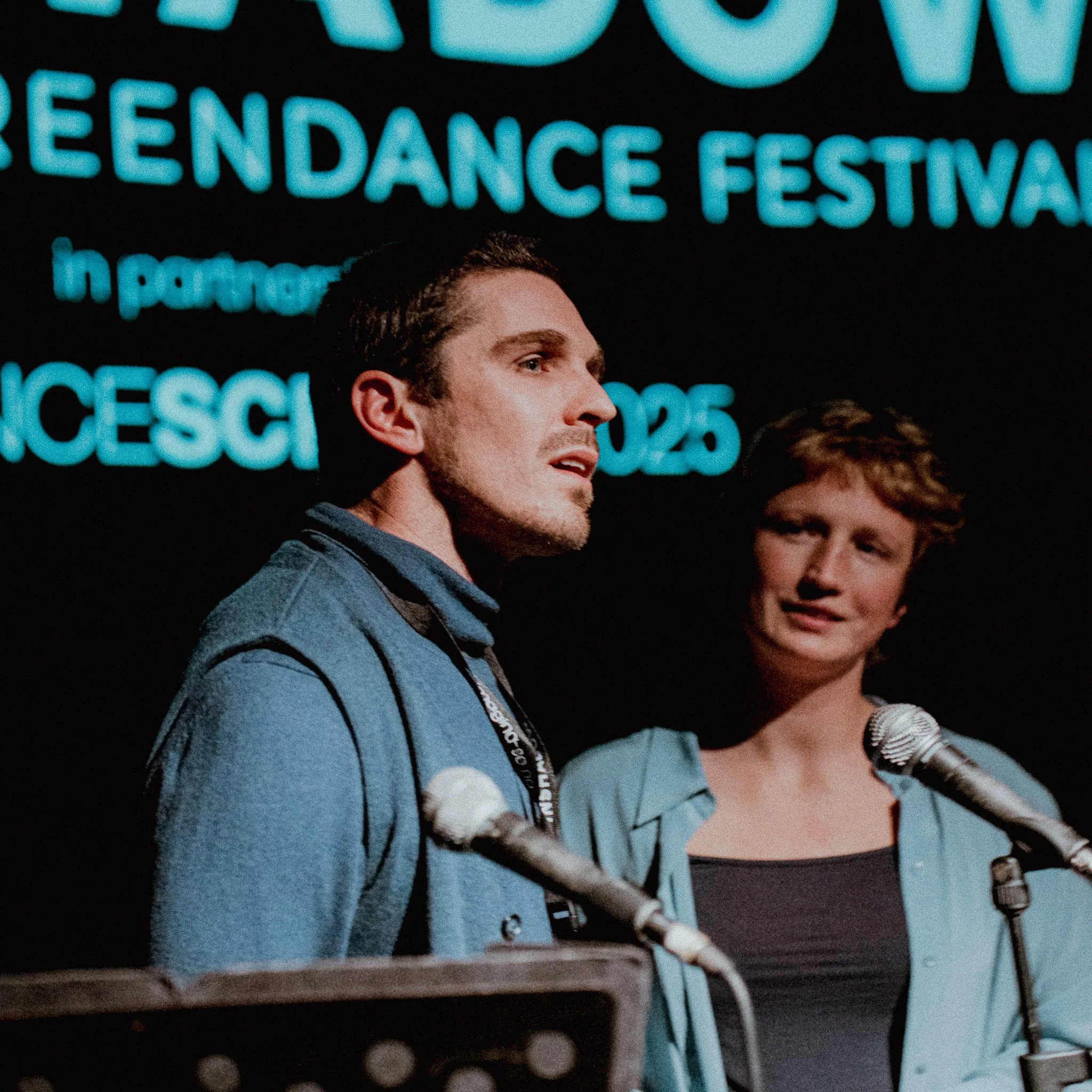Close-up of a person with brown hair and blue eyes, wearing a blue patterned jacket, speaking into a silver microphone at a public event or conference, with a large illuminated sign in the background that reads 'ENDORSEMENT FESTIVAL'.