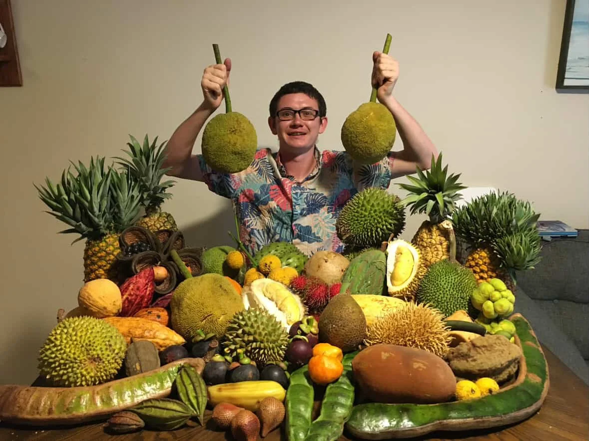 Xain of Xains World Nursery is positioned behind a pile of exotic fruit, holding up  two large and round fruits, possibly jackfruit