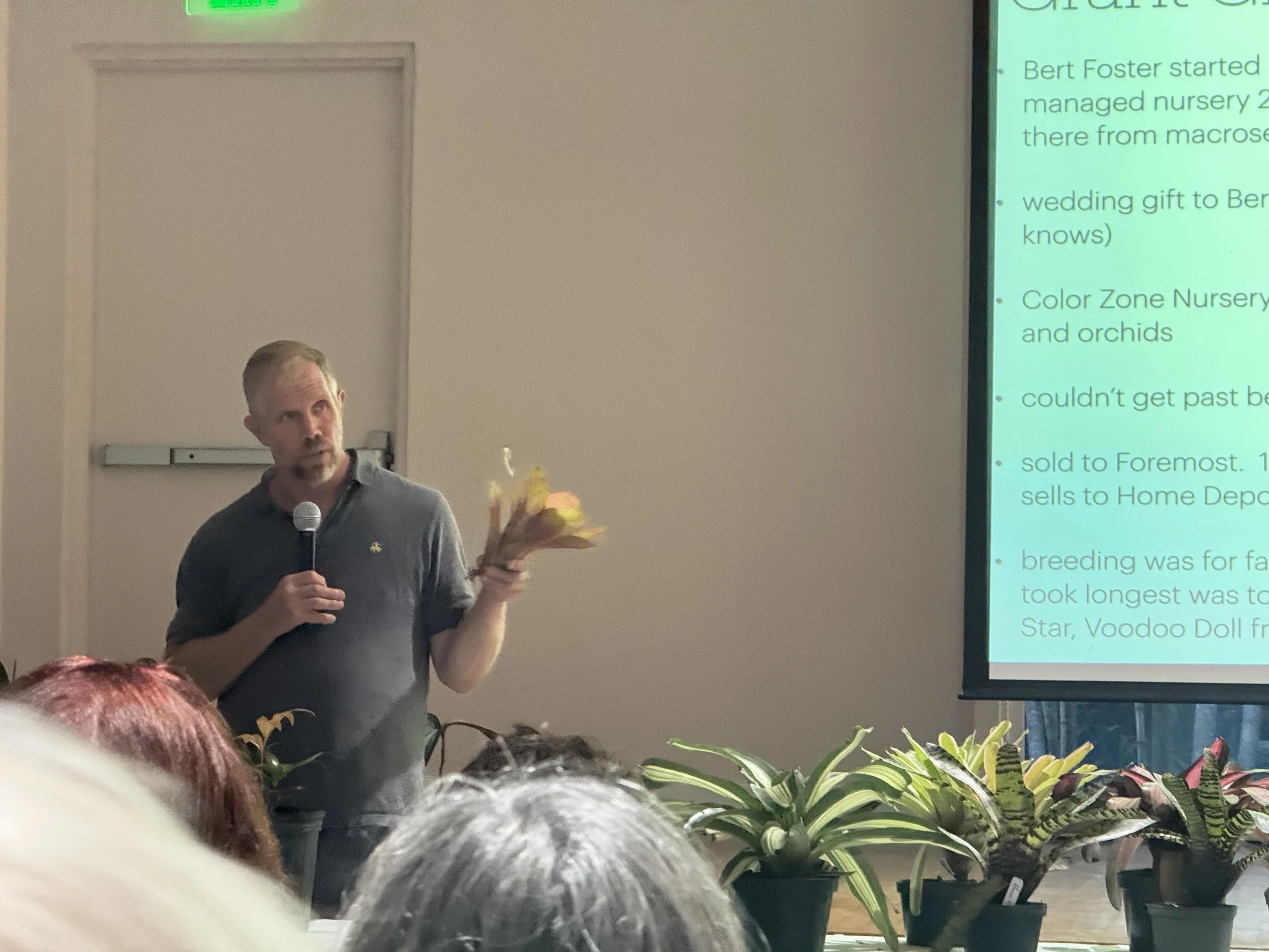 Chip Jones holding a bromeliad and speaking into a microphone in front of a presentation slide with interview questions asked to Bert Forster cut off