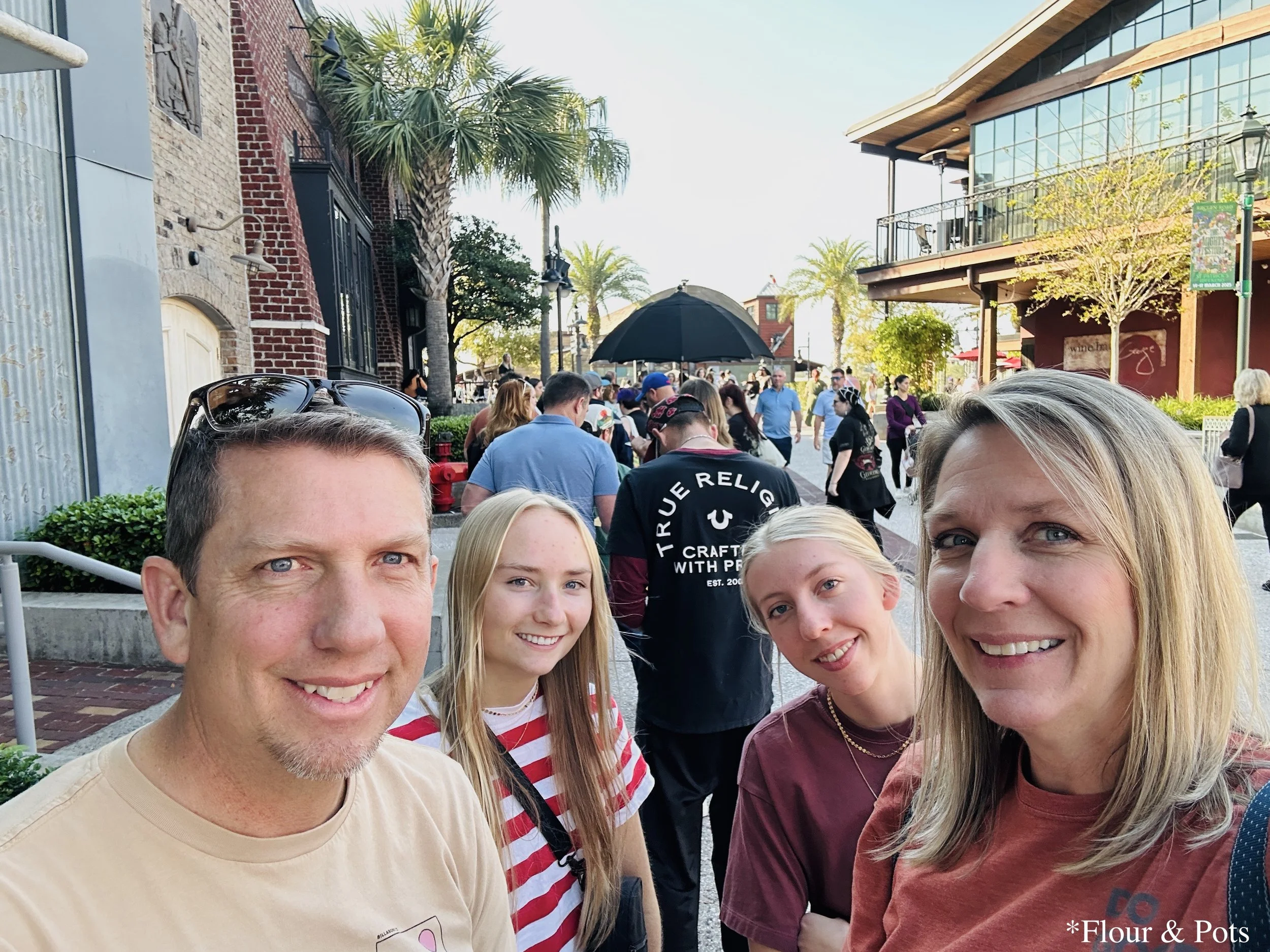 Family waiting in line outside Gideon's Bakehouse at Disney Springs on a sunny afternoon, surrounded by other visitors eager to try the famous cookies.