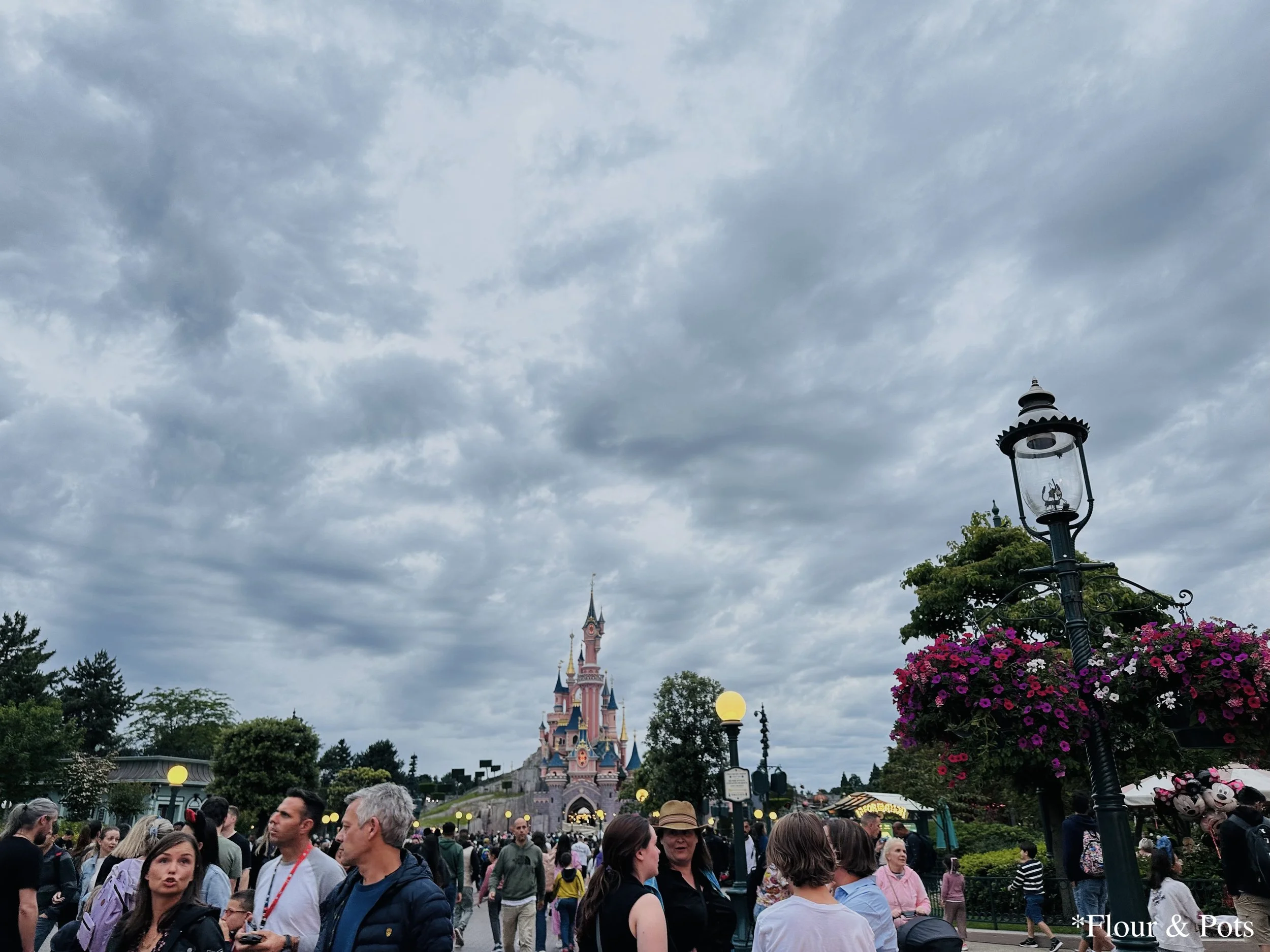 Sleeping Beauty’s Castle at Disneyland Paris glowing in the early evening light, with stormy gray clouds gathering overhead for a dramatic sky backdrop.