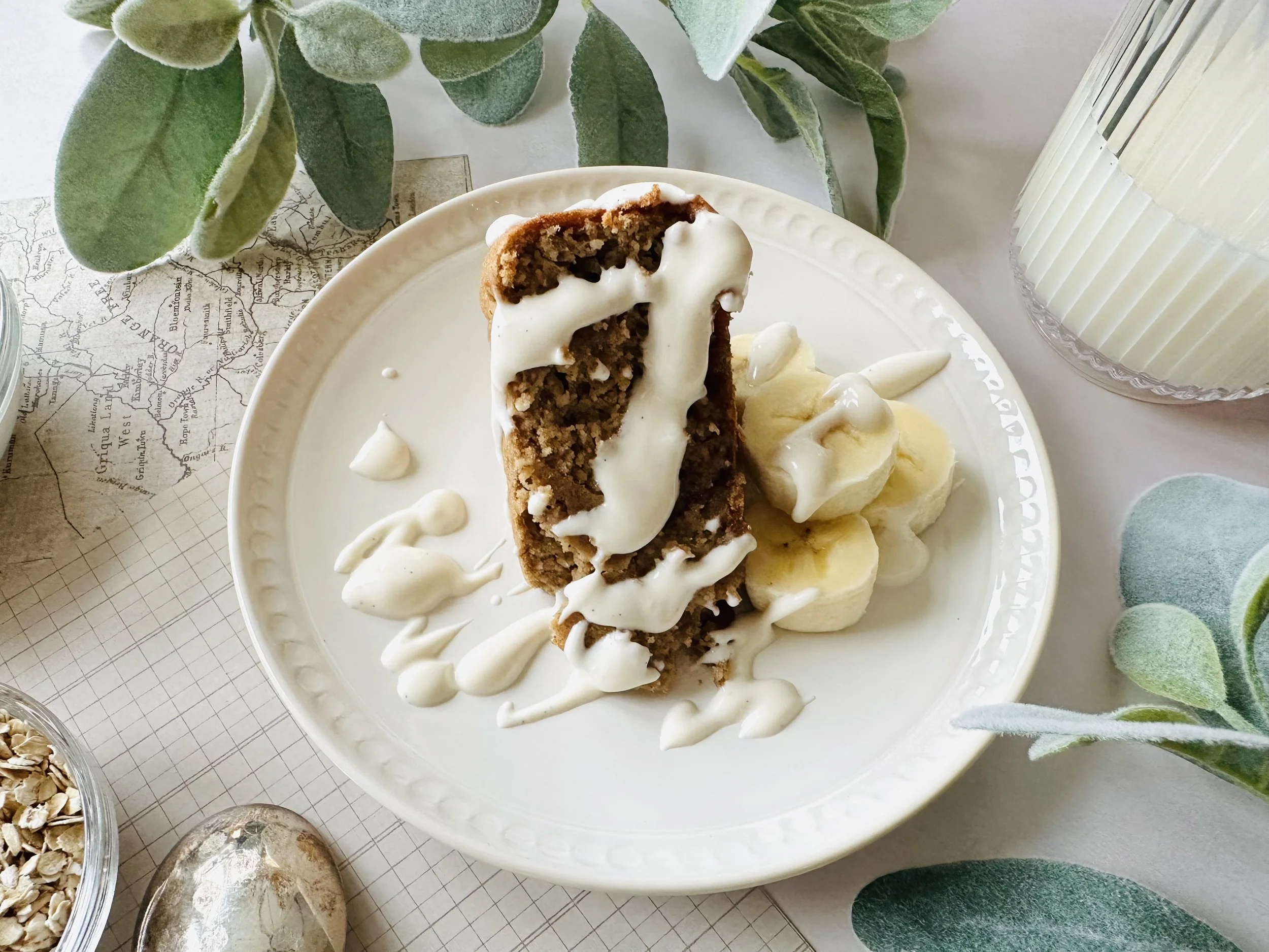 A slice of healthier blended oatmeal cake baked and topped with a greek yogurt dressing, garnished with banana slices and surrounded by green plants, a glass of milk, a bowl of oats, and a spoon to dig right into this delicious breakfast recipe.