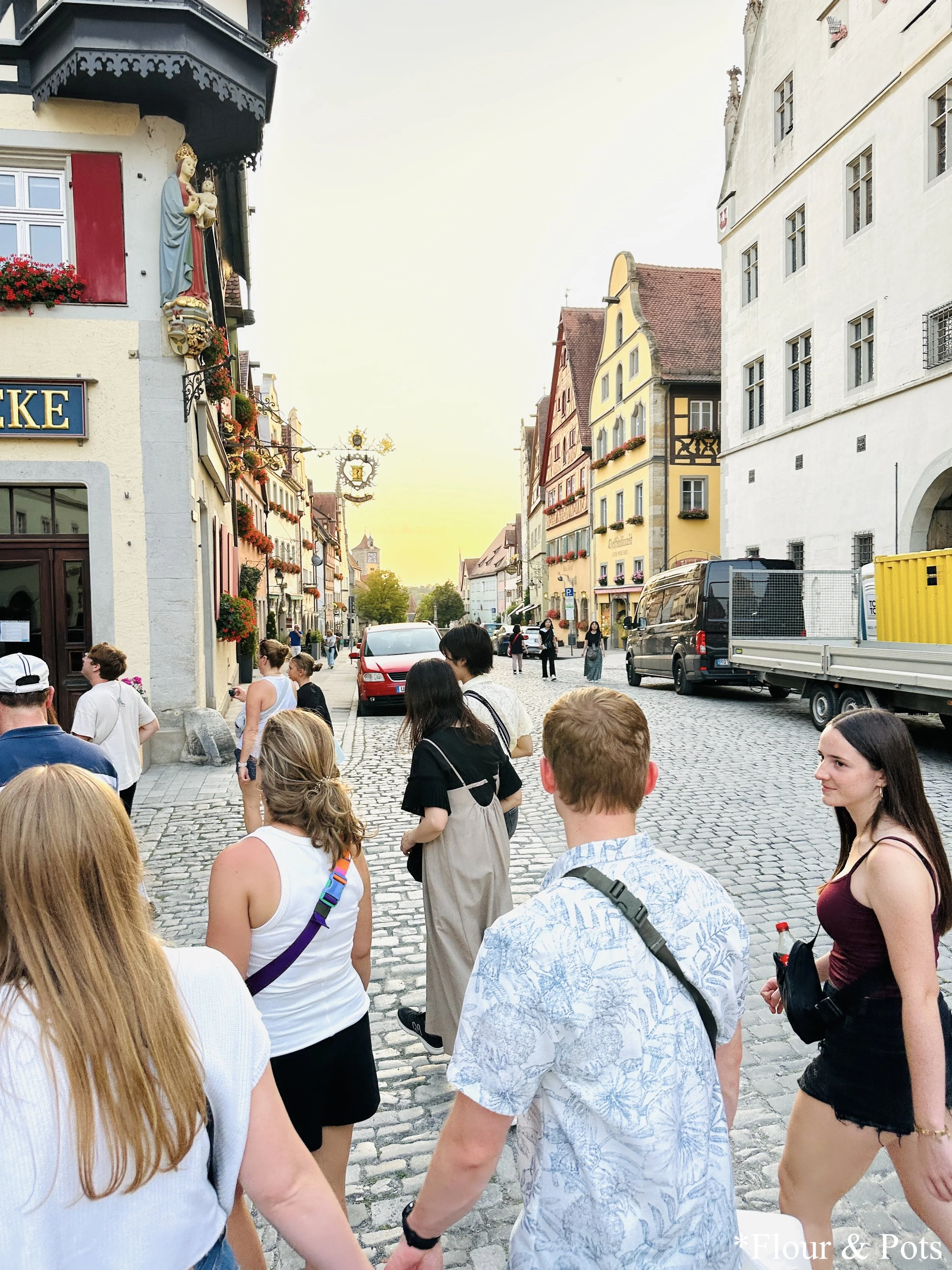 A group of tourists listening to the Night Watchman during his evening tour in Rothenburg, Germany, with the town's historic buildings and medieval city walls in the background.