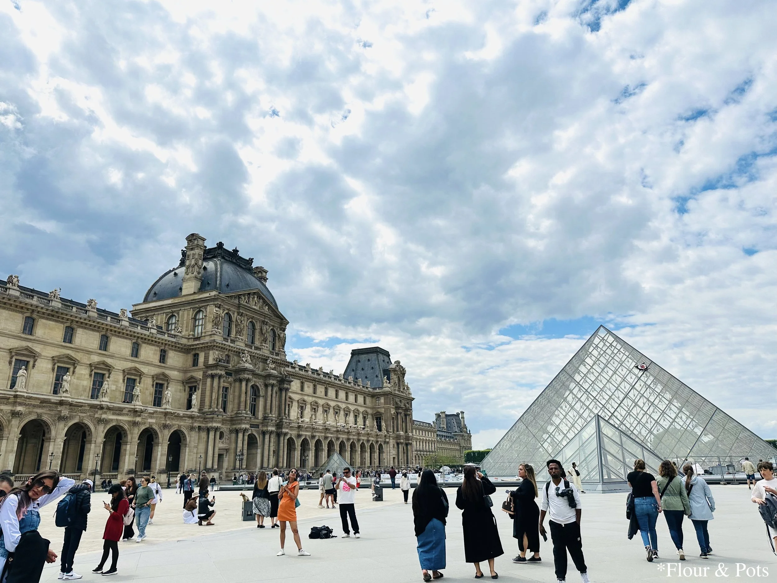 The iconic Louvre Palace and its glass pyramid bustling with tourists on a bright day in Paris, France.