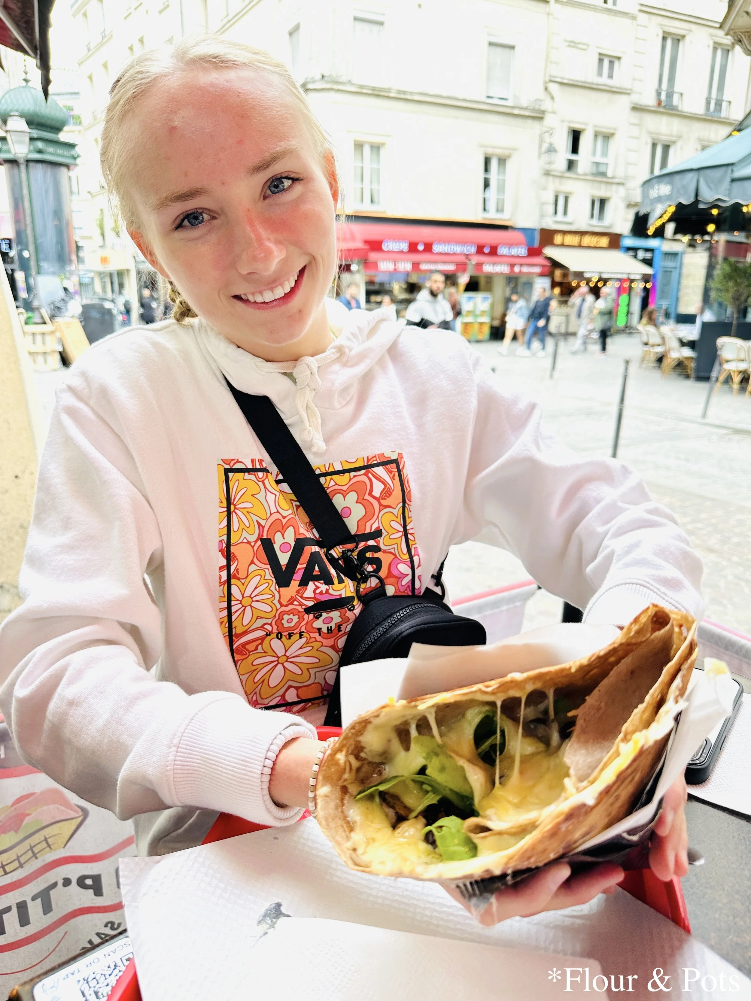 Me savoring a warm, cheesy crêpe from Au P’tit Grec Crêperie on a lively Paris street, with the café and city scene in the background.