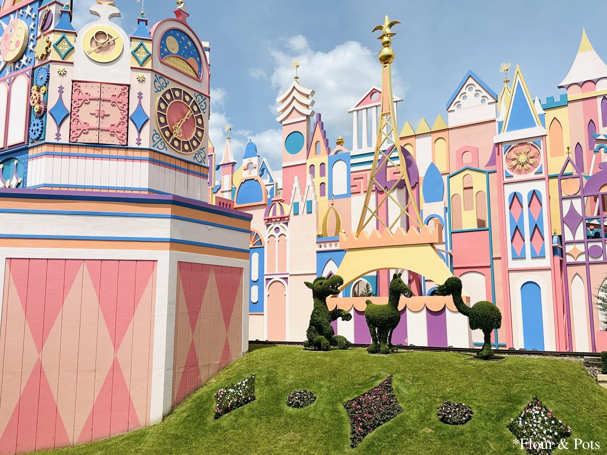right pastel-colored exterior of It’s a Small World at Disneyland Paris, with decorative flower beds and animal-shaped trimmed bushes on the sloped lawn in front of the attraction.