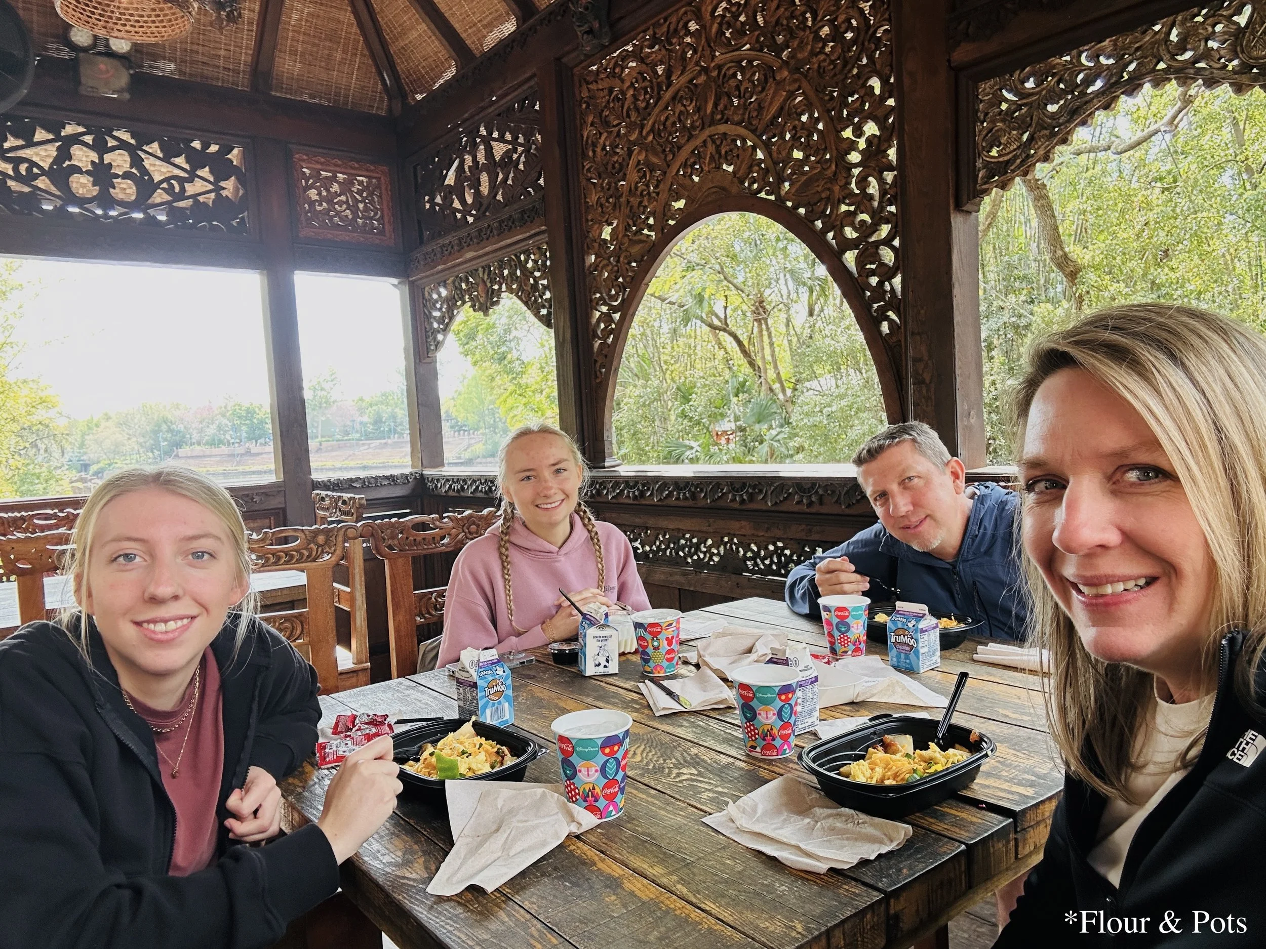 Family enjoying breakfast from Yak & Yeti Local Food Café while seated at tables near the main river in the center of Disney's Animal Kingdom, Orlando, Florida, surrounded by lush greenery and a peaceful park atmosphere.