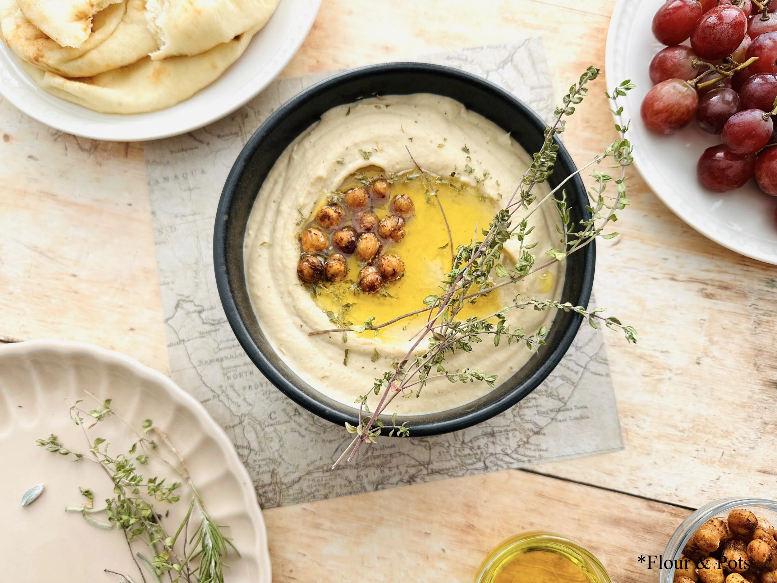 A bowl of creamy hummus made with Greek yogurt and white beans, surrounded by pita bread, fresh grapes, herbs, a drizzle of olive oil, and chickpeas, creating a vibrant and inviting snack spread.