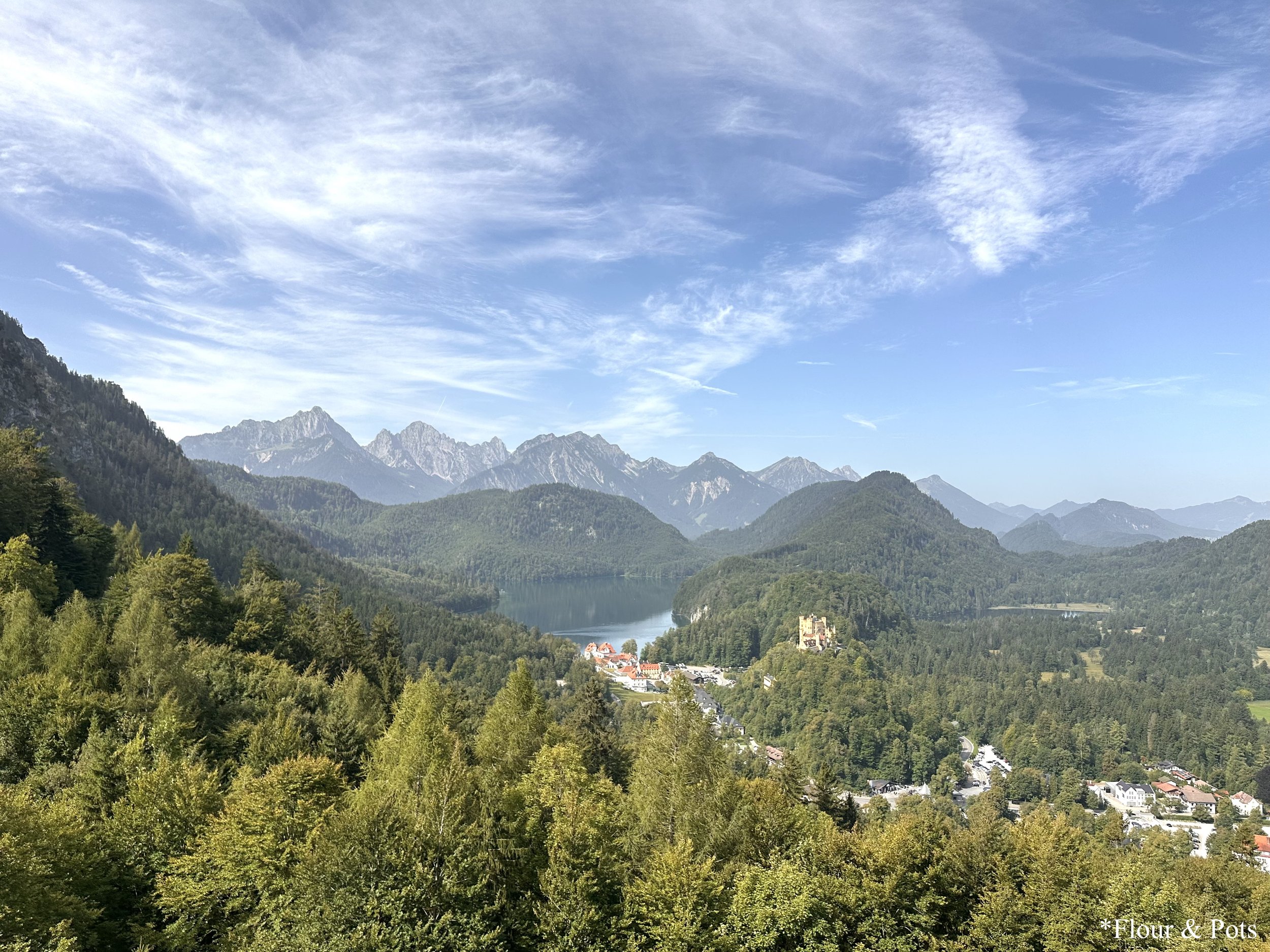 Scenic view of Hohenschwangau Castle overlooking the Alpsee lake, with lush green mountains in the background.