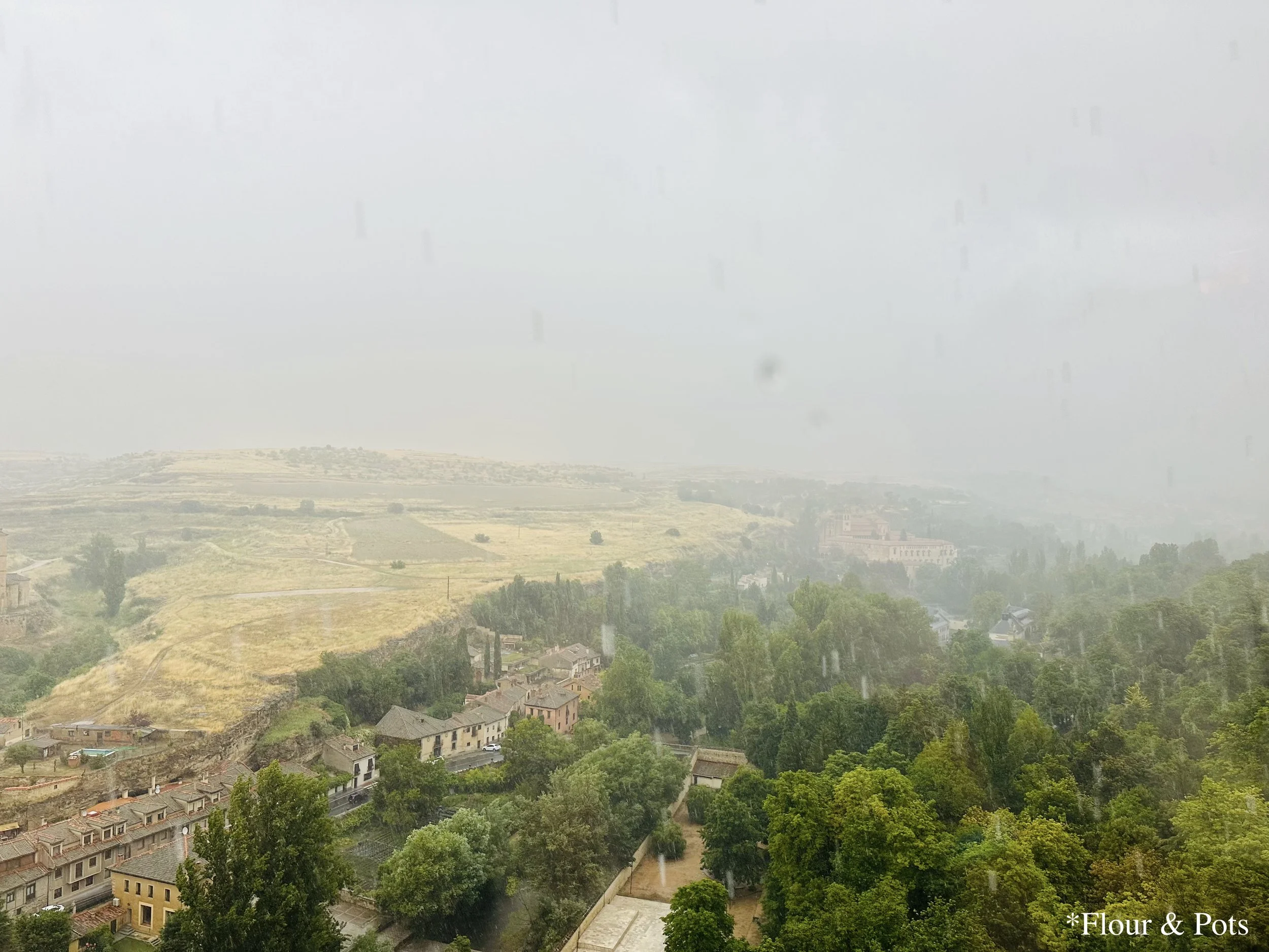 A view of rain falling from the castle’s covered walkway lookouts in Segovia, Spain. The stone arches frame the scene, with droplets of rain visible as they cascade down the stone walls.