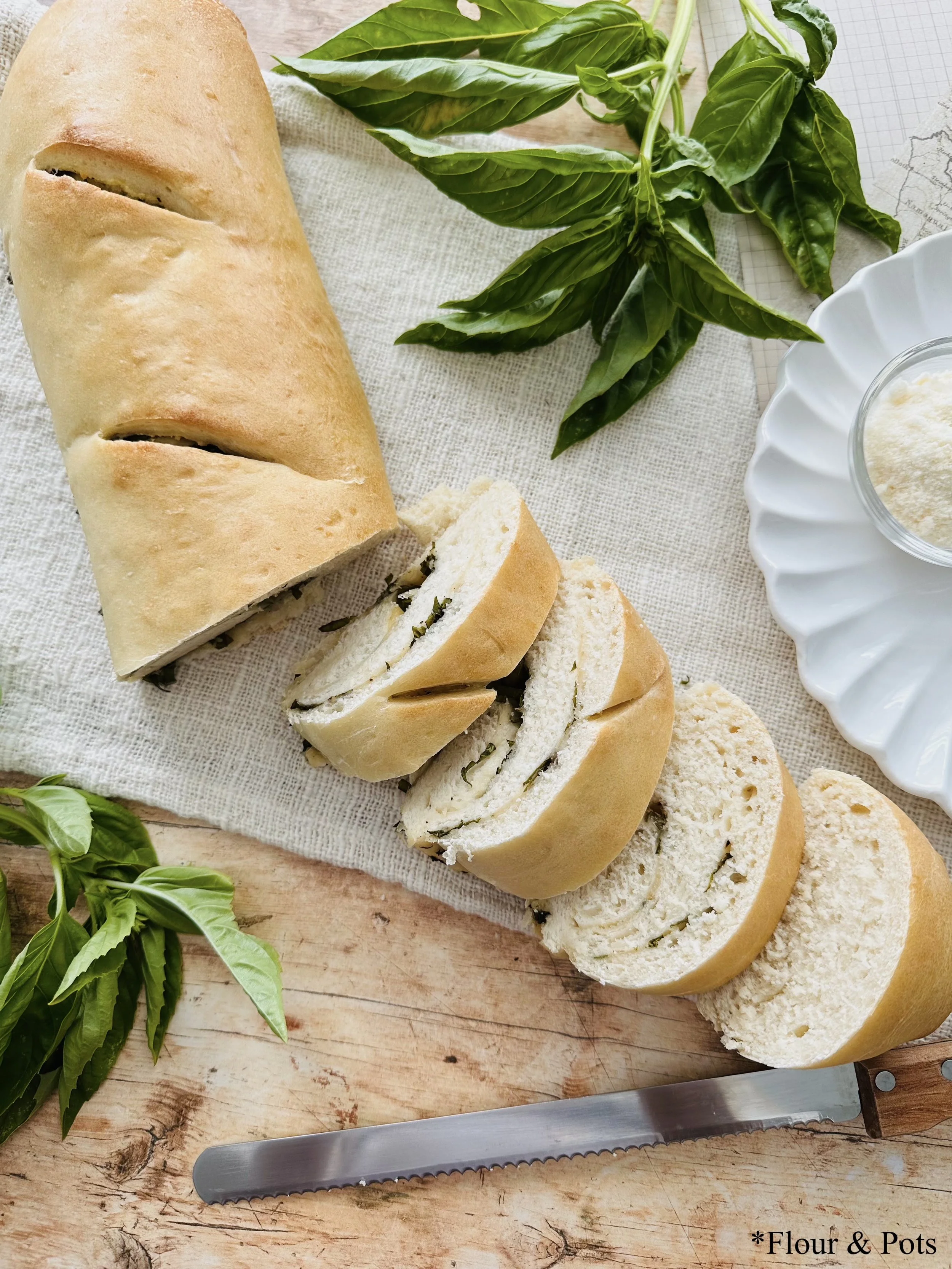 A full loaf of Cacio e Pepe French bread, half sliced, resting on a woven towel with fresh basil leaves off to the side.