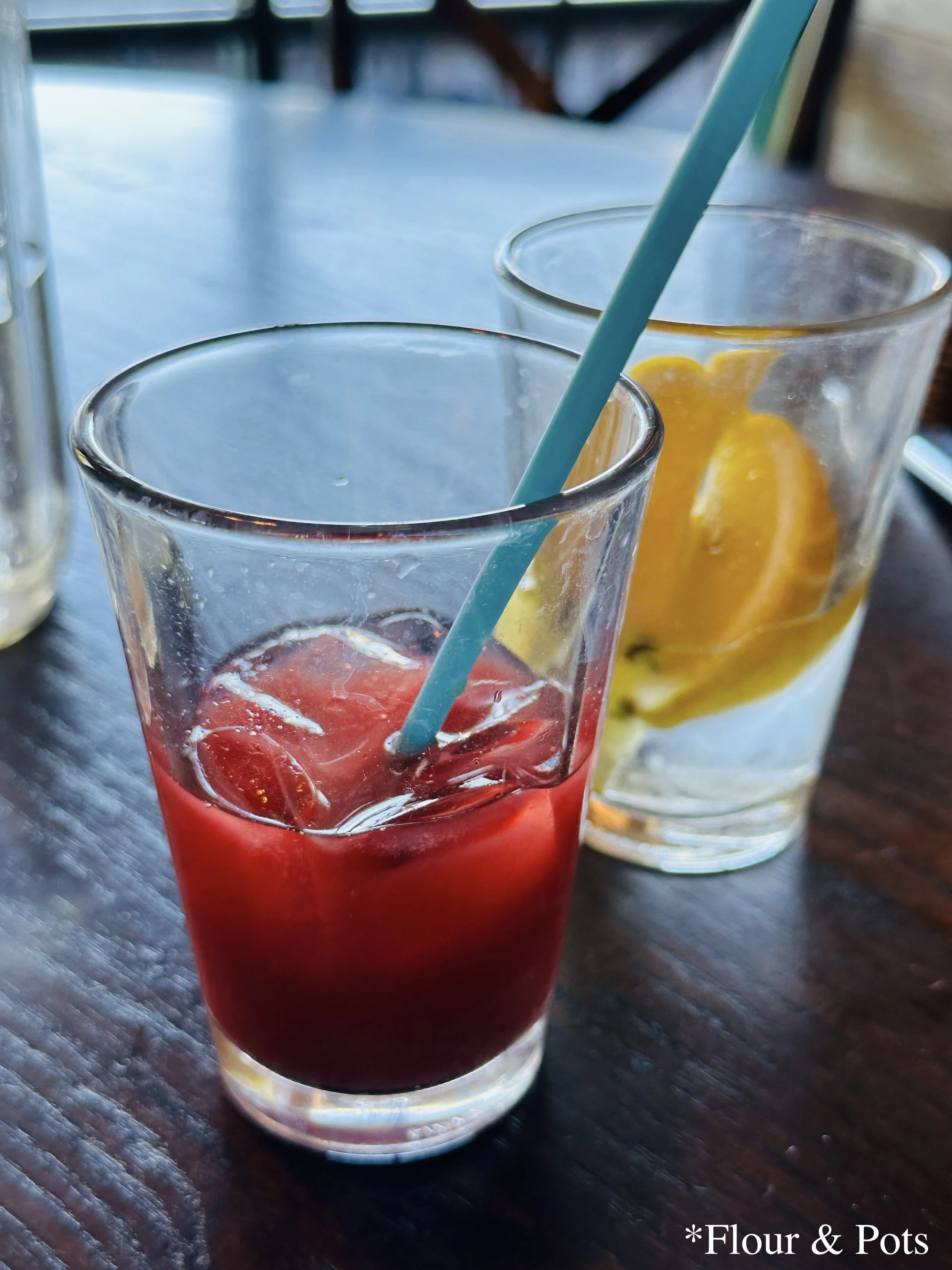 Two vibrant, healthy juices from First Watch on a breakfast table — the orange-hued Morning Meditation Fresh Juice and the deep purple seasonal Blue Booster Juice, served in clear glasses, ready to be enjoyed.