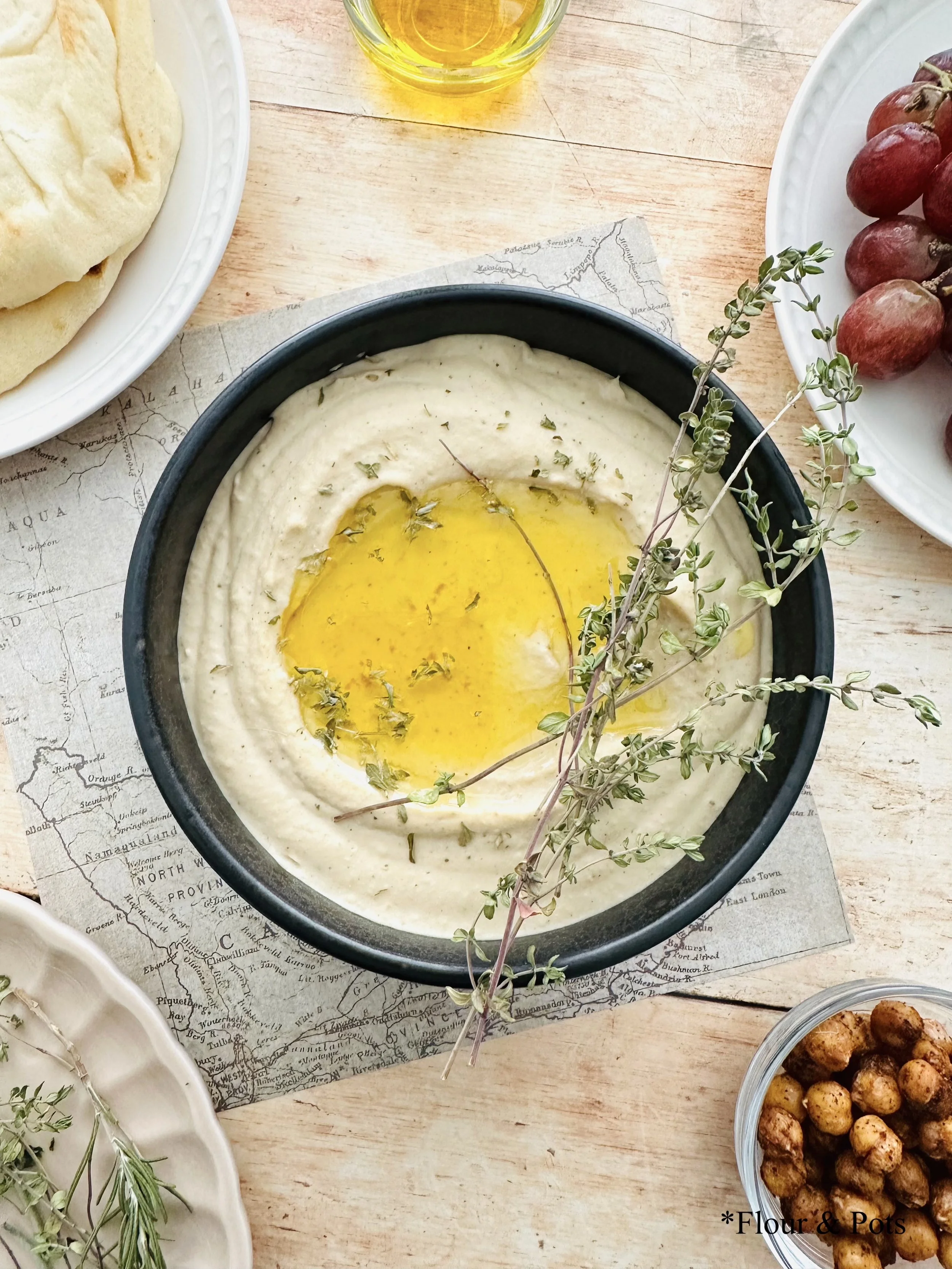 Horizontal image of creamy hummus made with Greek yogurt and white beans, surrounded by pita bread, fresh grapes, herbs, olive oil, and roasted chickpeas, creating a vibrant, inviting spread.