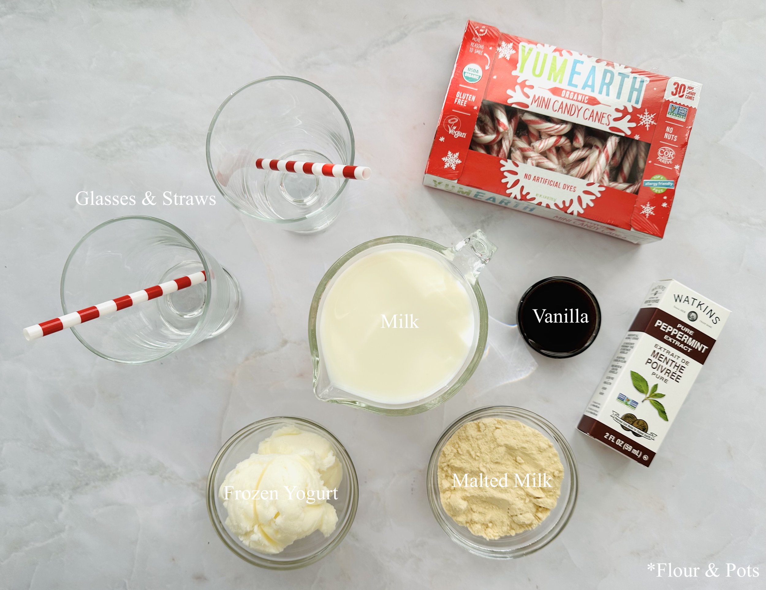 Assorted ingredients for peppermint candy cane smoothies arranged on a light surface, including milk, frozen yogurt, malted milk powder, and candy canes.